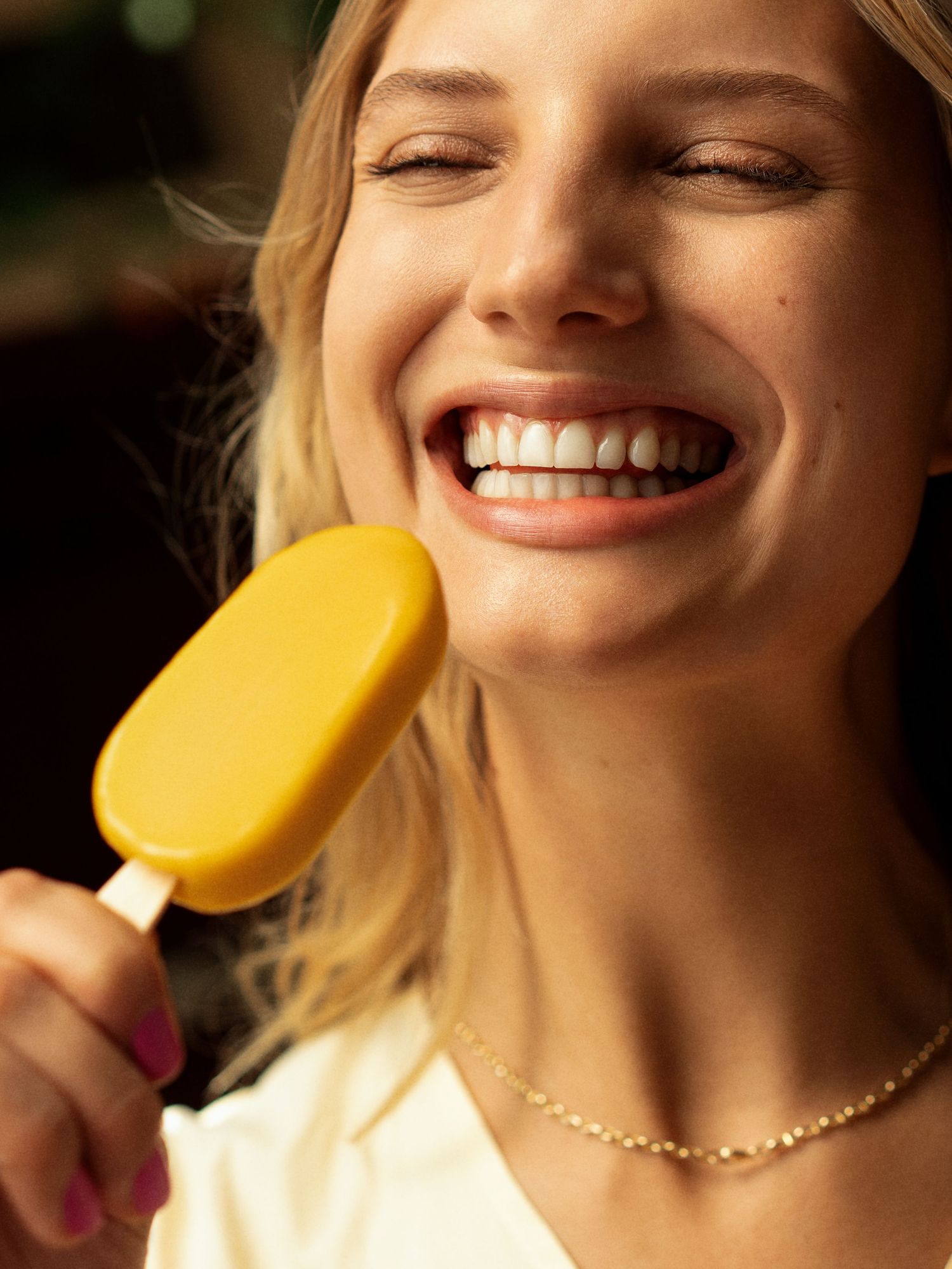 Happy blonde woman laughing while eating a yellow mango ice cream popsicle outdoors.