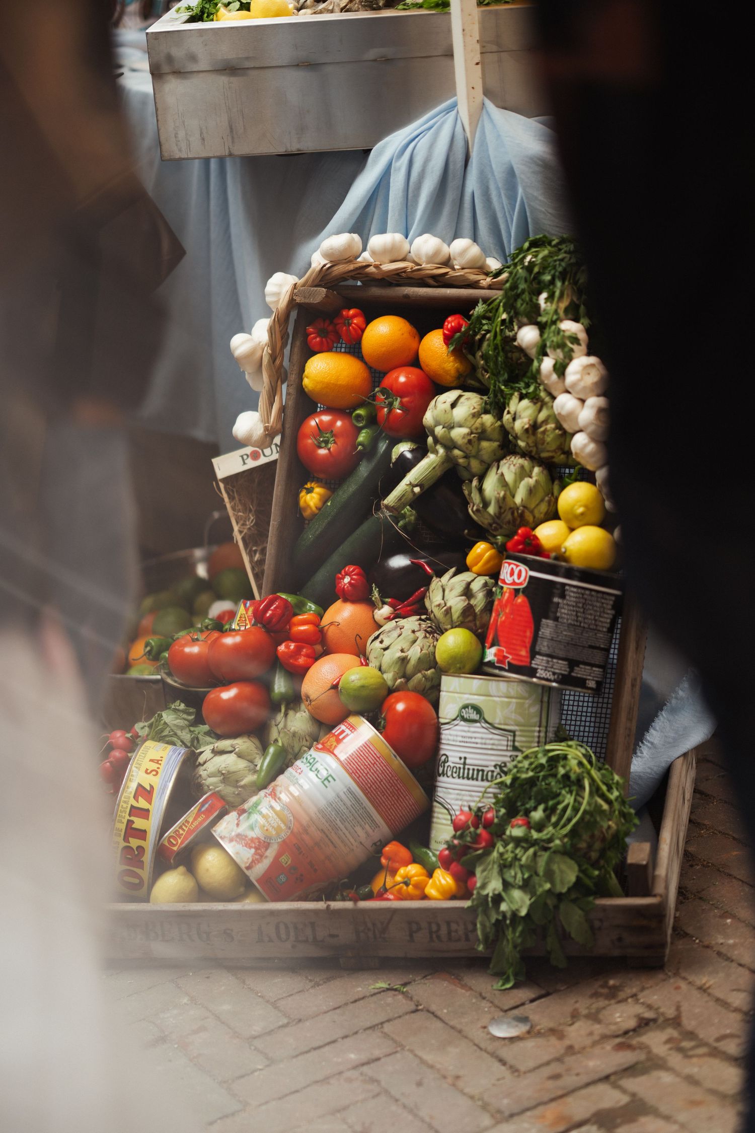 Fresh vegetables, fruits, and canned goods arranged in wooden crates at a busy outdoor farmers market.
