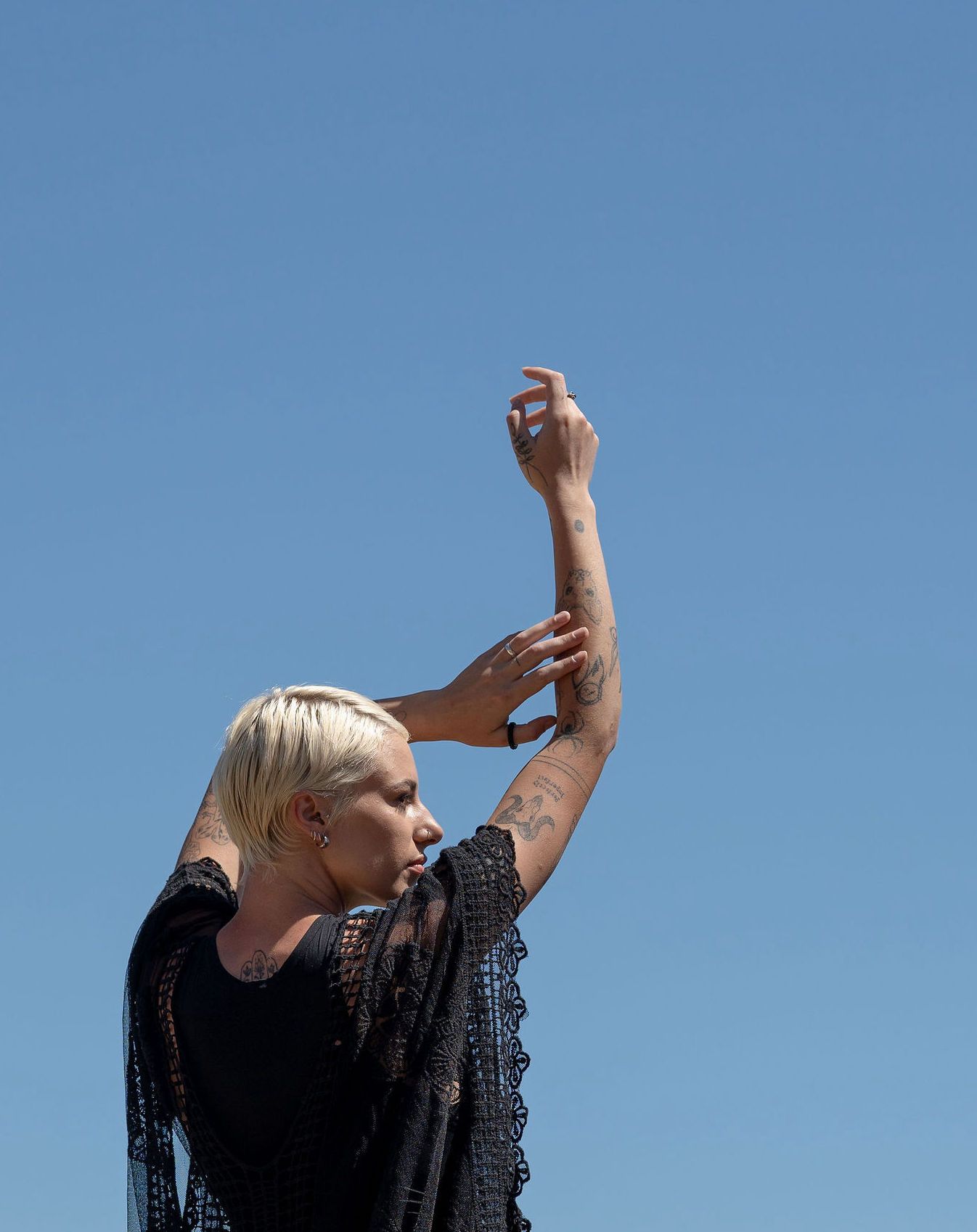 Woman with blonde hair in black lace top raises arm gracefully against clear blue sky.