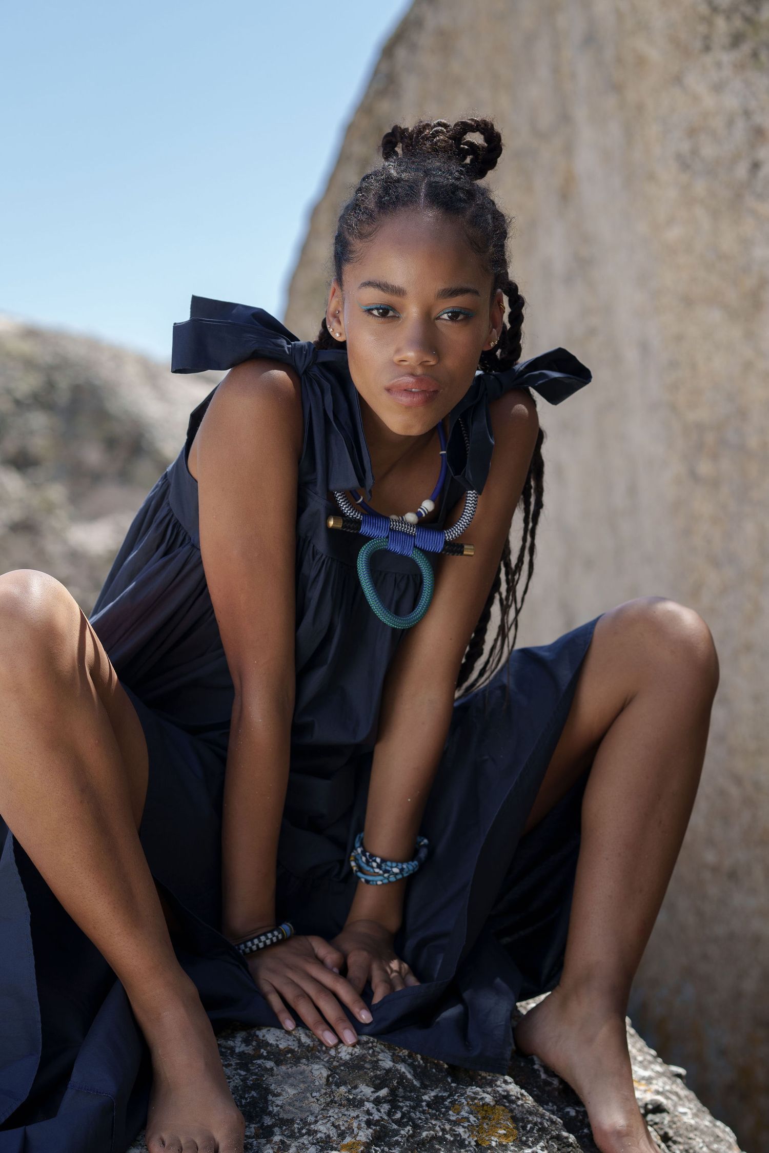 Young Black woman in navy dress with braids sits on rocks outdoors, wearing colorful necklace and bracelets.
