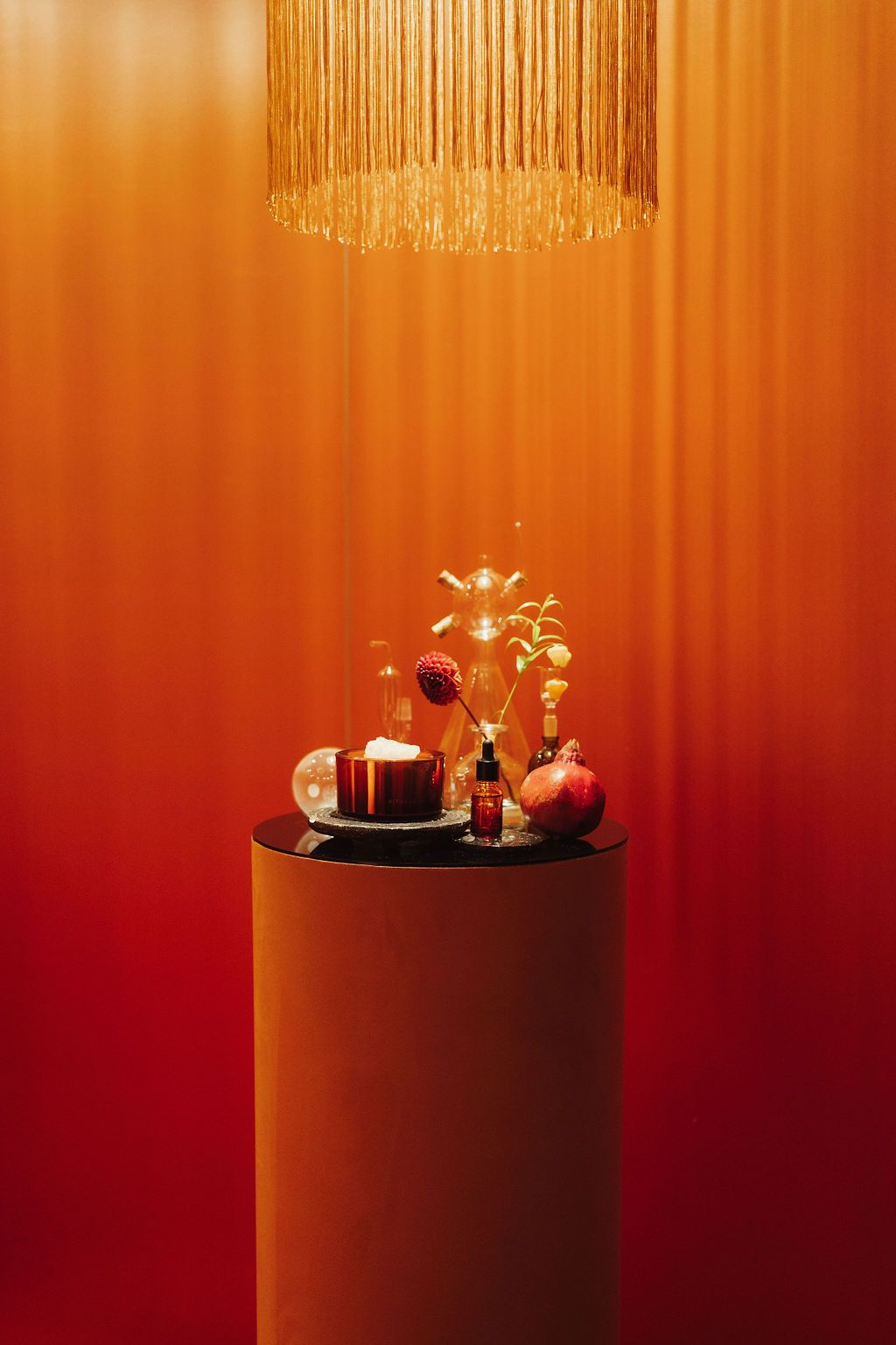 Elegant display on dark pedestal with candle, botanicals, and pomegranate under gold fringe lamp against orange backdrop.