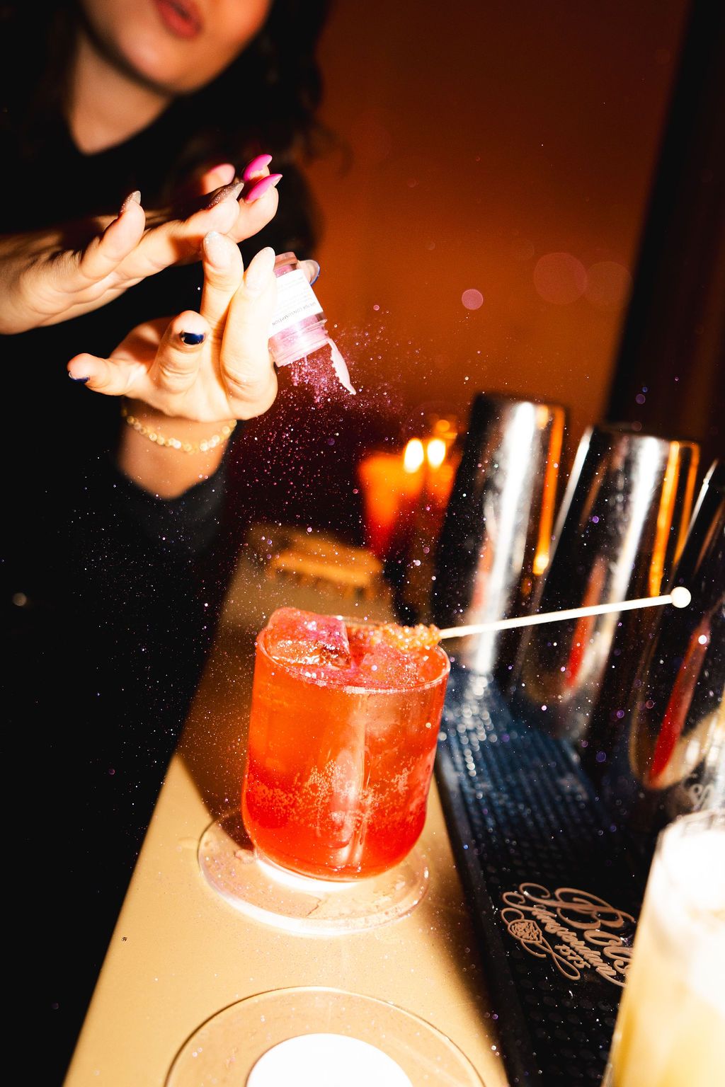 Bartender sprinkling glitter on a red cocktail in a dark bar setting with cocktail shakers nearby.