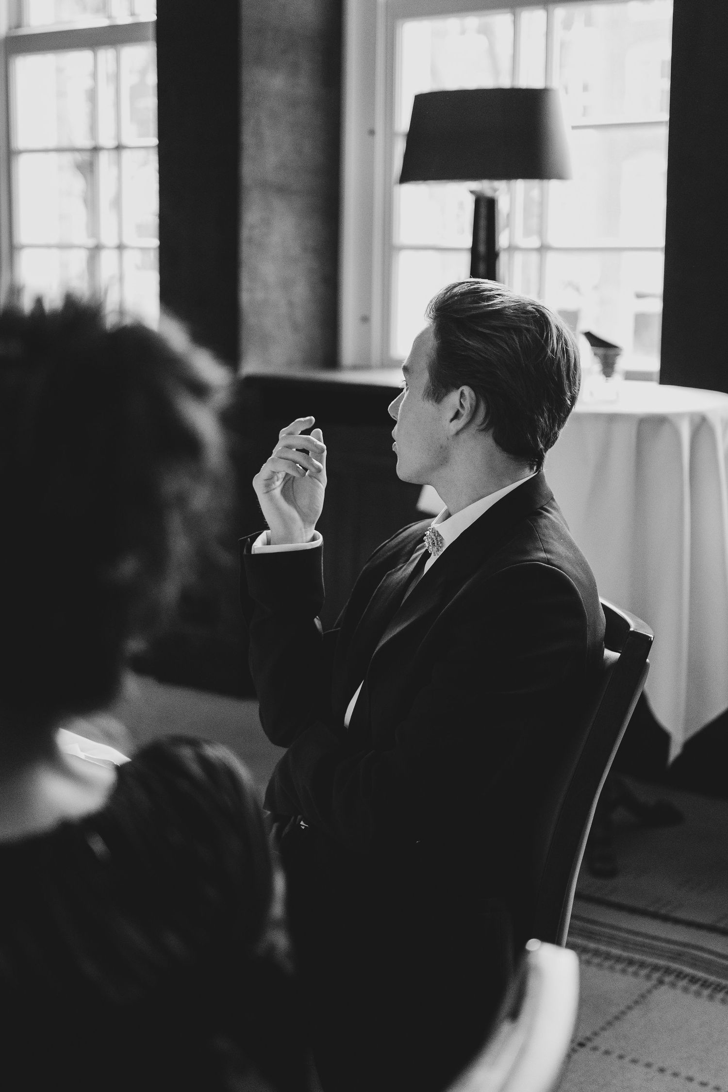 Black and white photo of a man in a suit sitting in a chair, holding a small object while speaking at a formal event.
