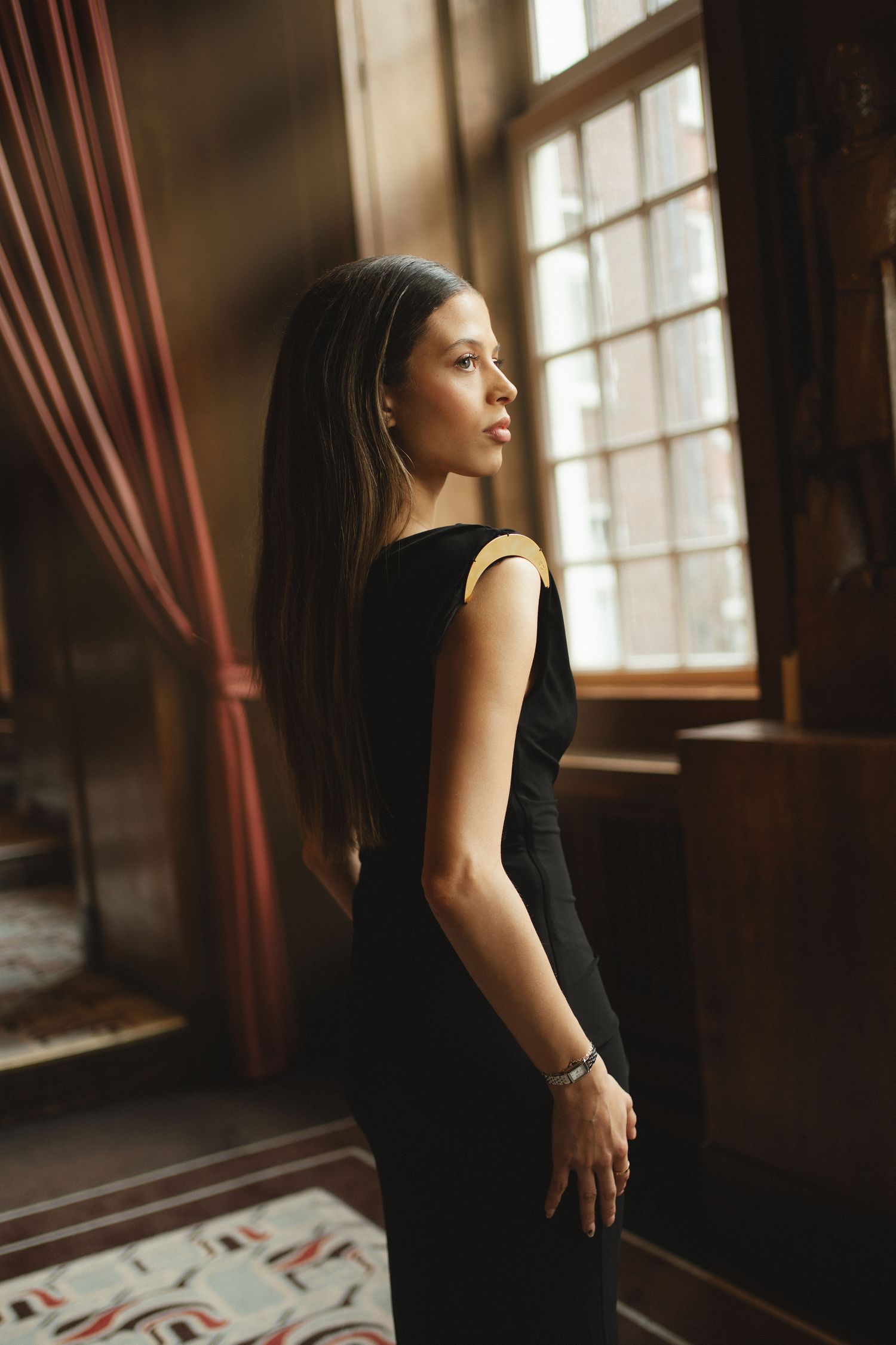 Elegant woman in black dress standing by vintage window in ornate interior, long hair, bracelet, soft natural light.