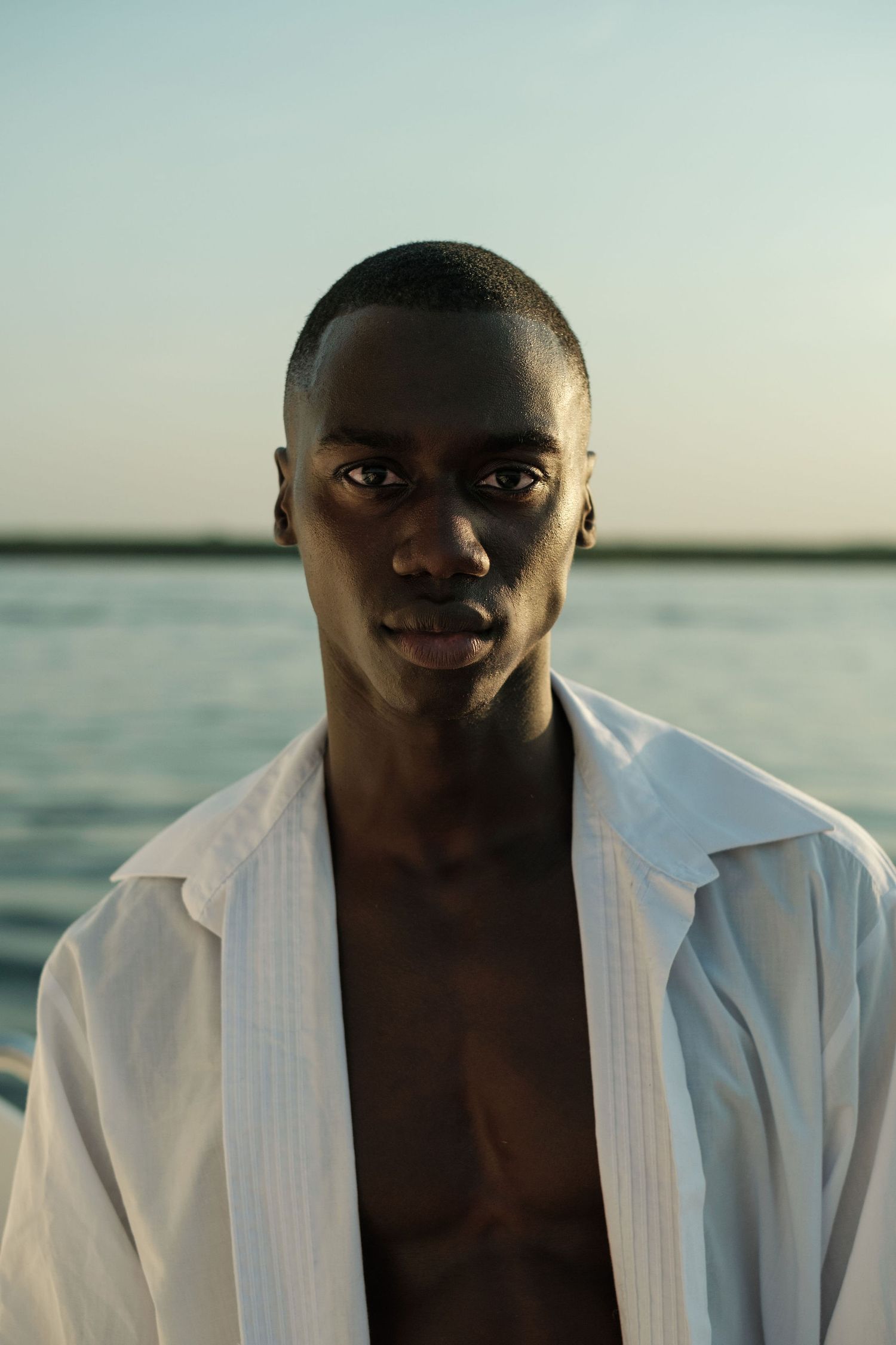 Young Black man in open white shirt poses confidently against a serene waterfront backdrop at dusk.