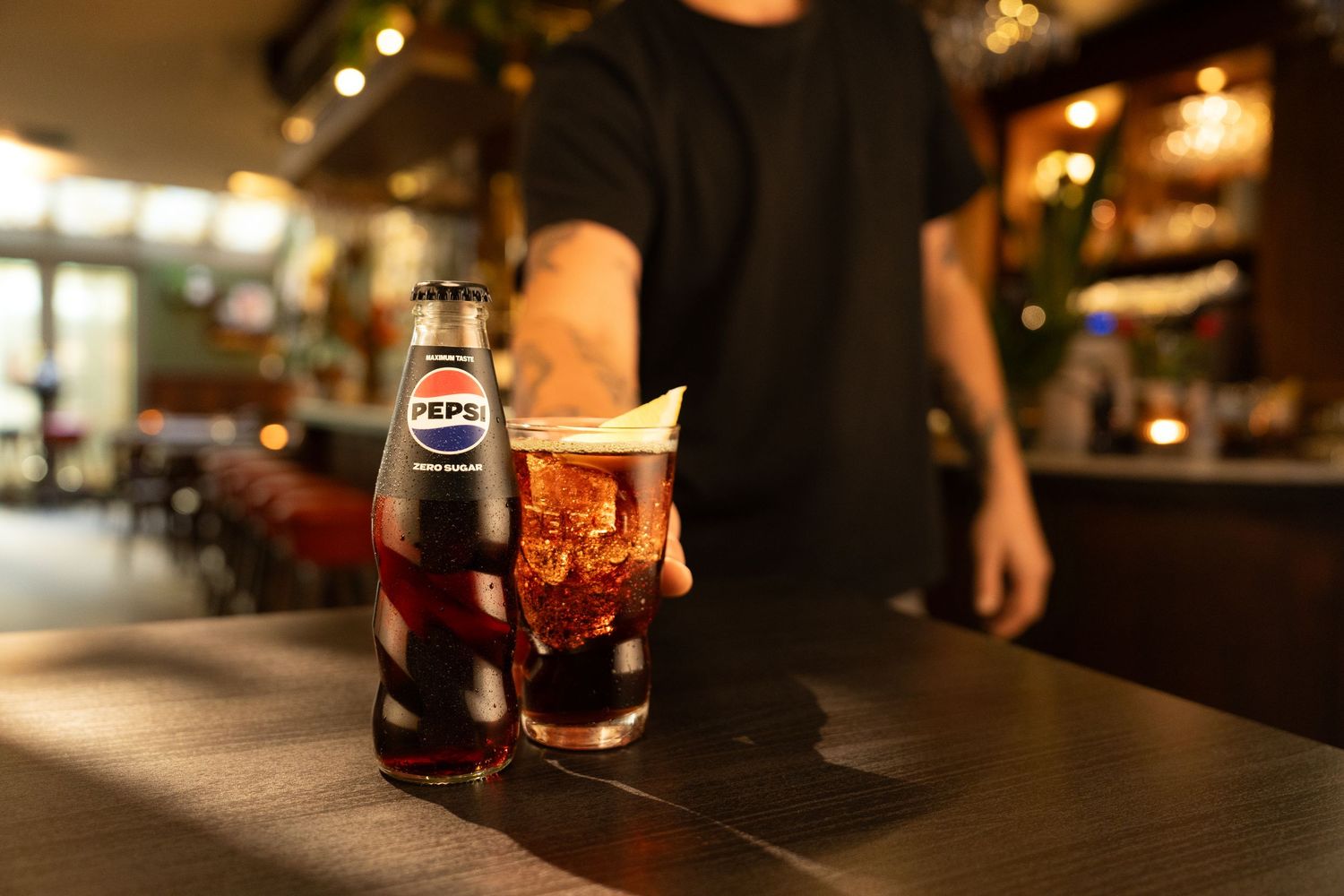A bartender serves a glass of Pepsi with ice and a Pepsi bottle on a dark bar counter in a restaurant.