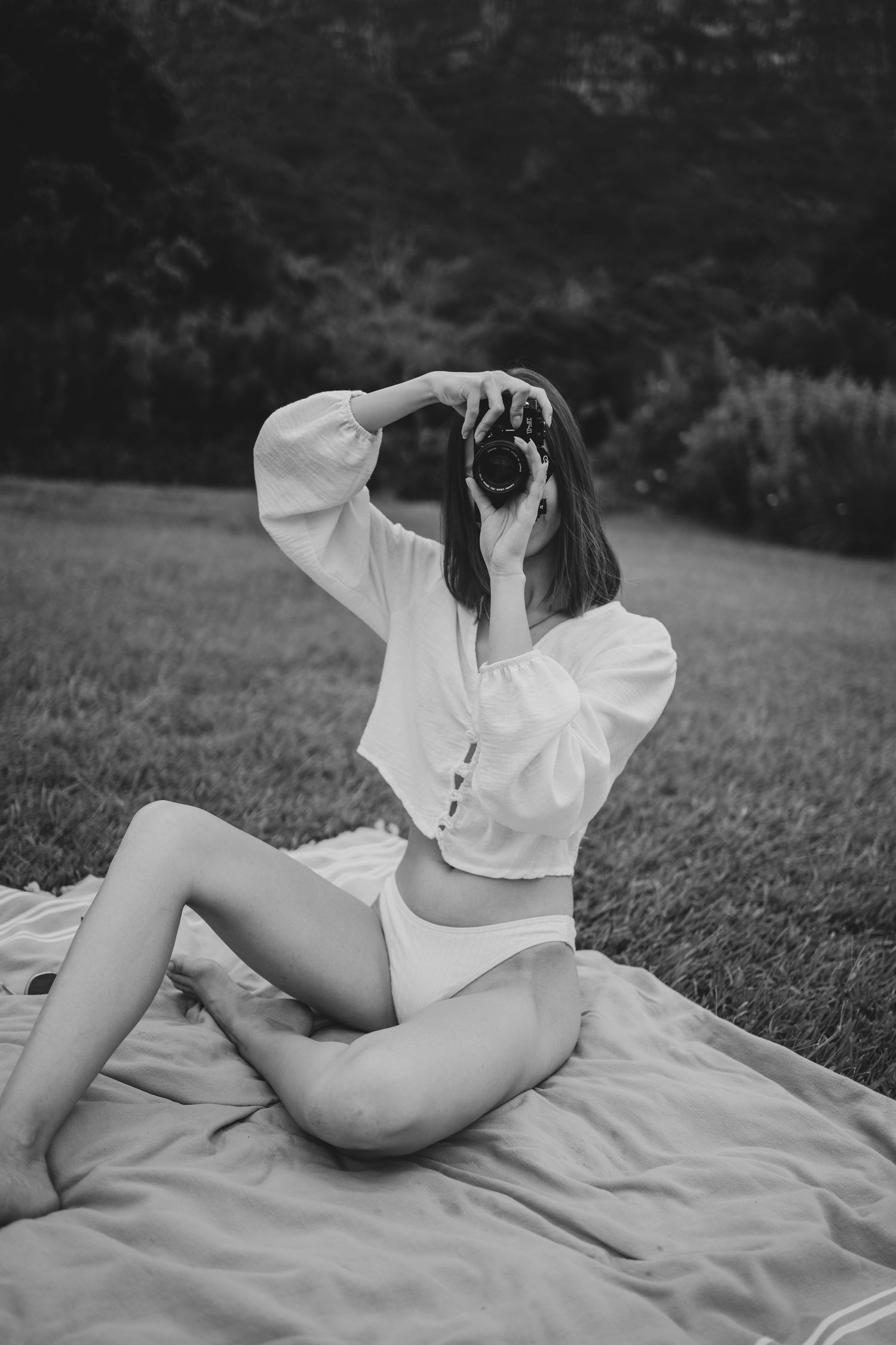 Black and white photo of a woman in white crop top and bikini bottoms sitting on a blanket outdoors holding a camera.