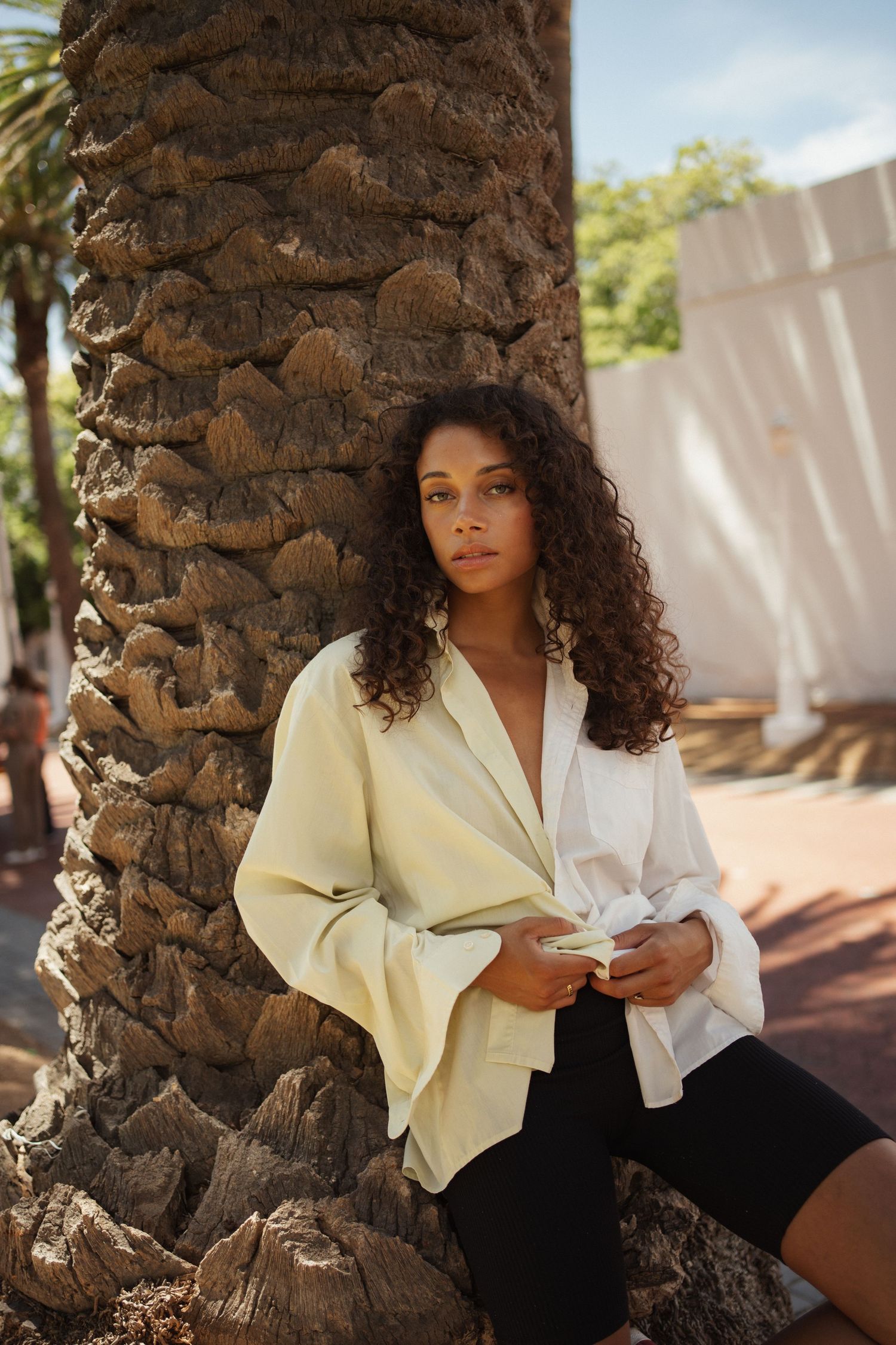 Young woman with curly hair in a two-tone white blazer leaning against a palm tree outdoors.