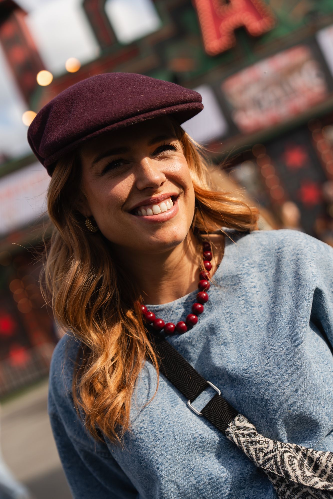 Smiling woman wearing a burgundy flat cap, red beaded necklace, and blue sweater outdoors at a festive venue.