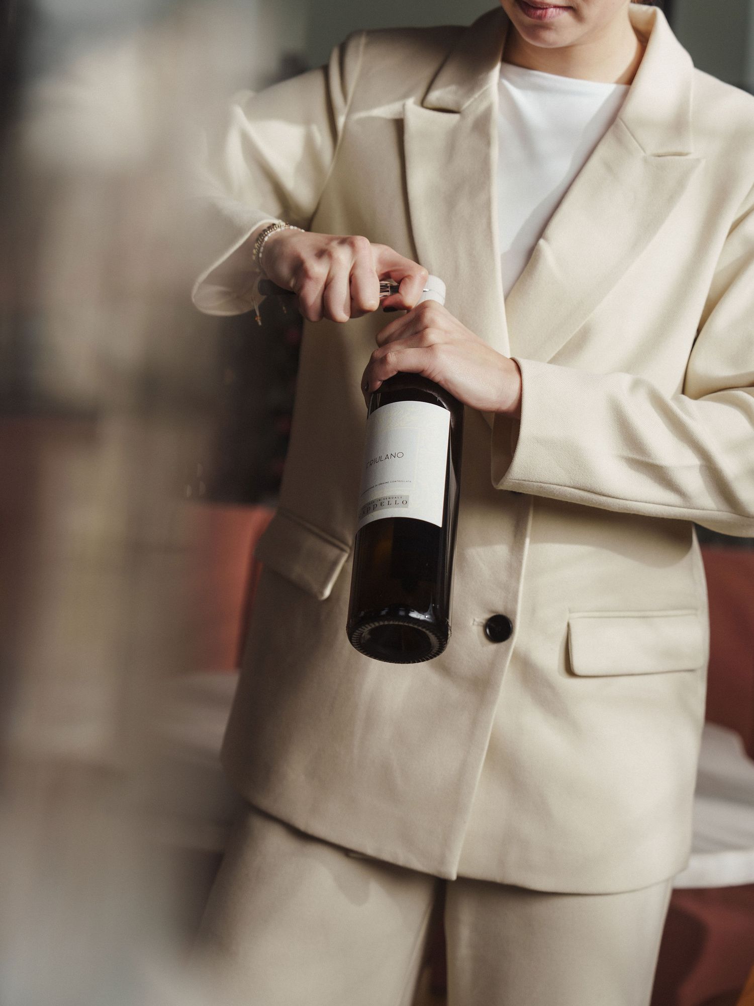 Person in beige suit opening a wine bottle, holding it carefully with both hands in an elegant setting.