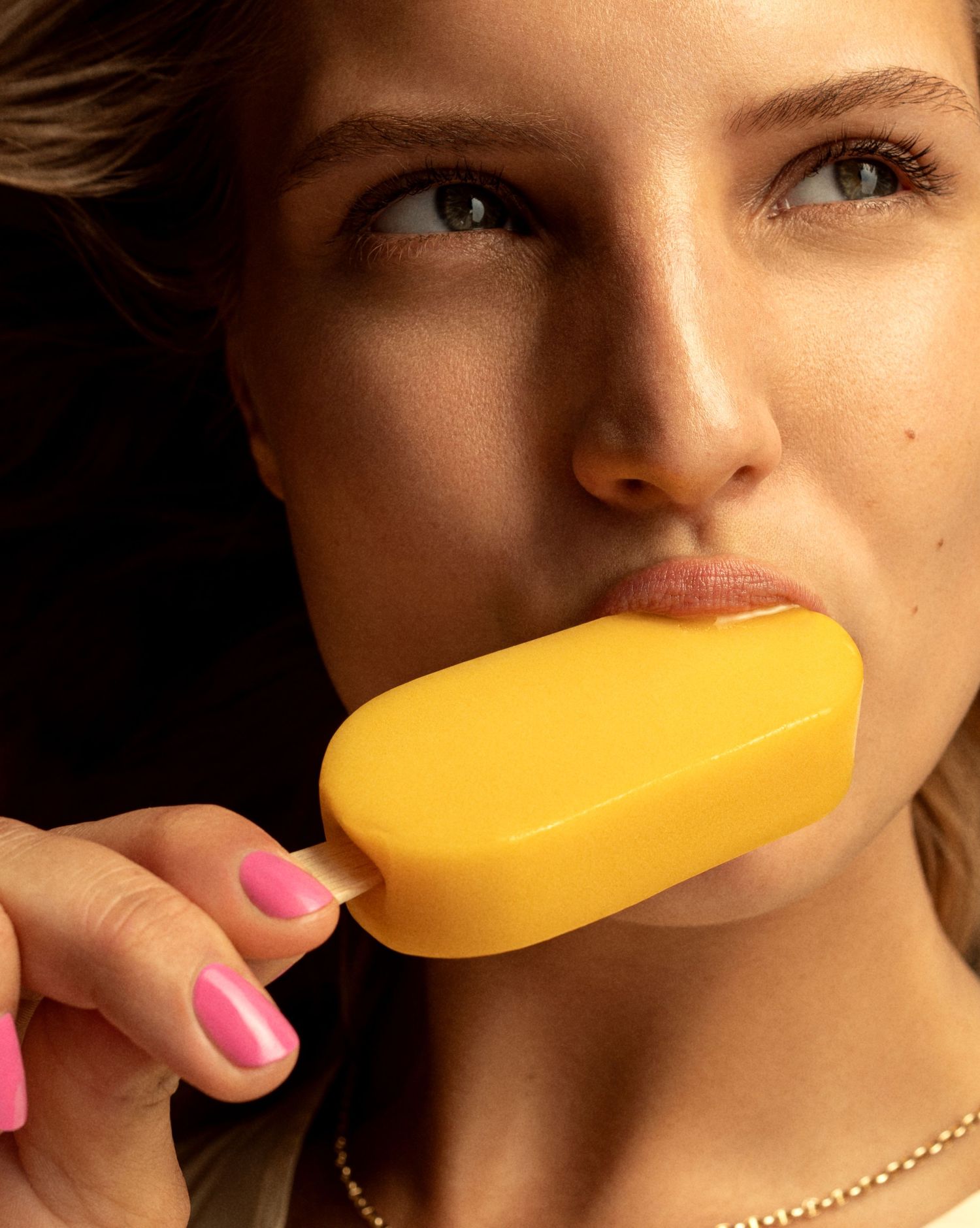 Woman sensually licking a yellow mango popsicle with pink nails against a dark background.