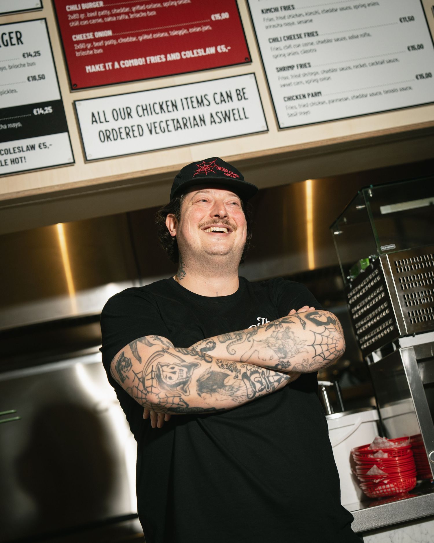 Tattooed chef in black cap smiling with crossed arms in front of a chicken restaurant menu board.