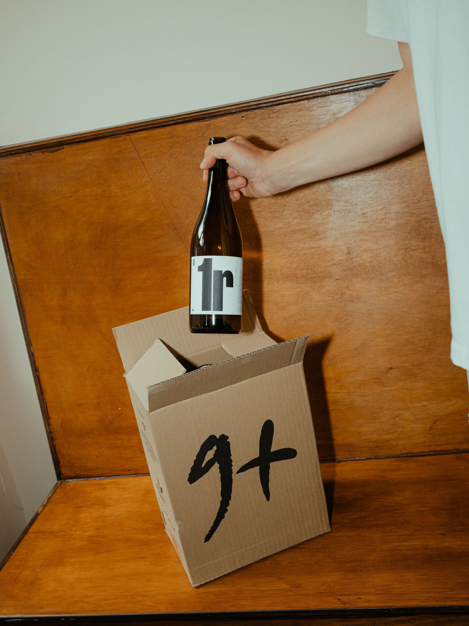 Hand pulling wine bottle labeled 1r from cardboard box marked 9+ on wooden bench.