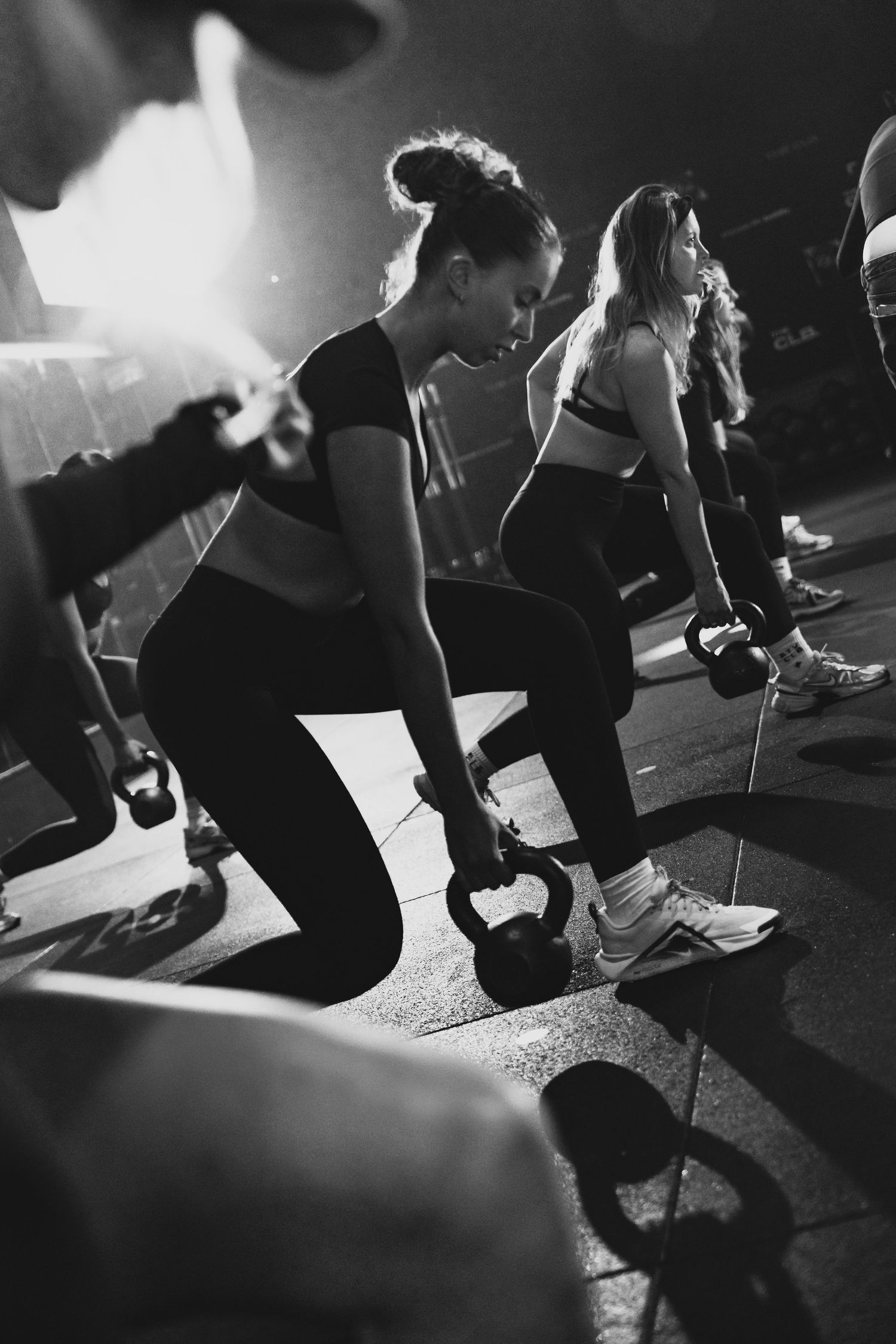 Women doing kettlebell lunges during a group fitness class in a dark gym, black and white photo.
