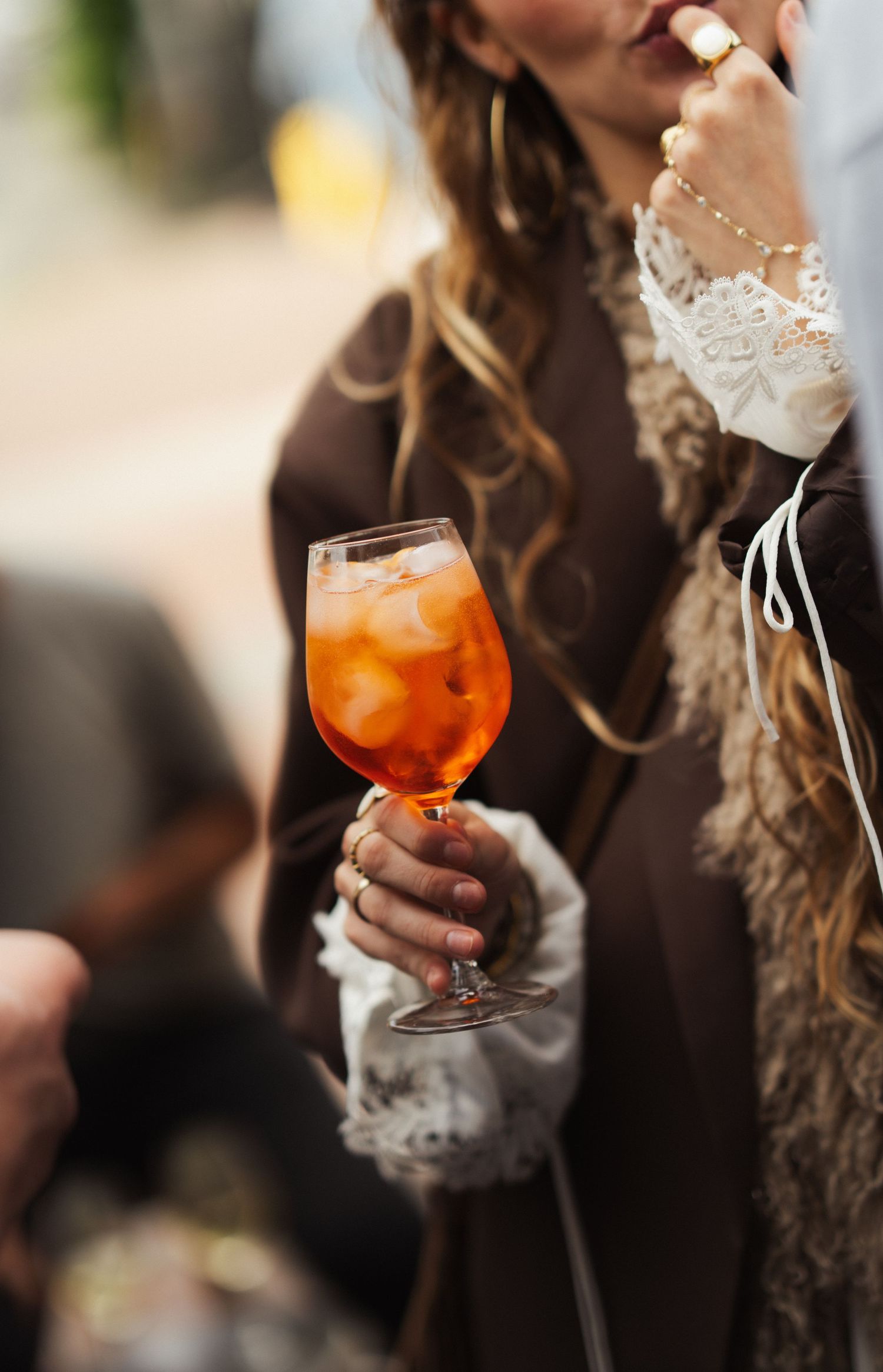 Woman holding an orange Aperol Spritz cocktail in a wine glass, wearing bohemian lace and fur fashion.