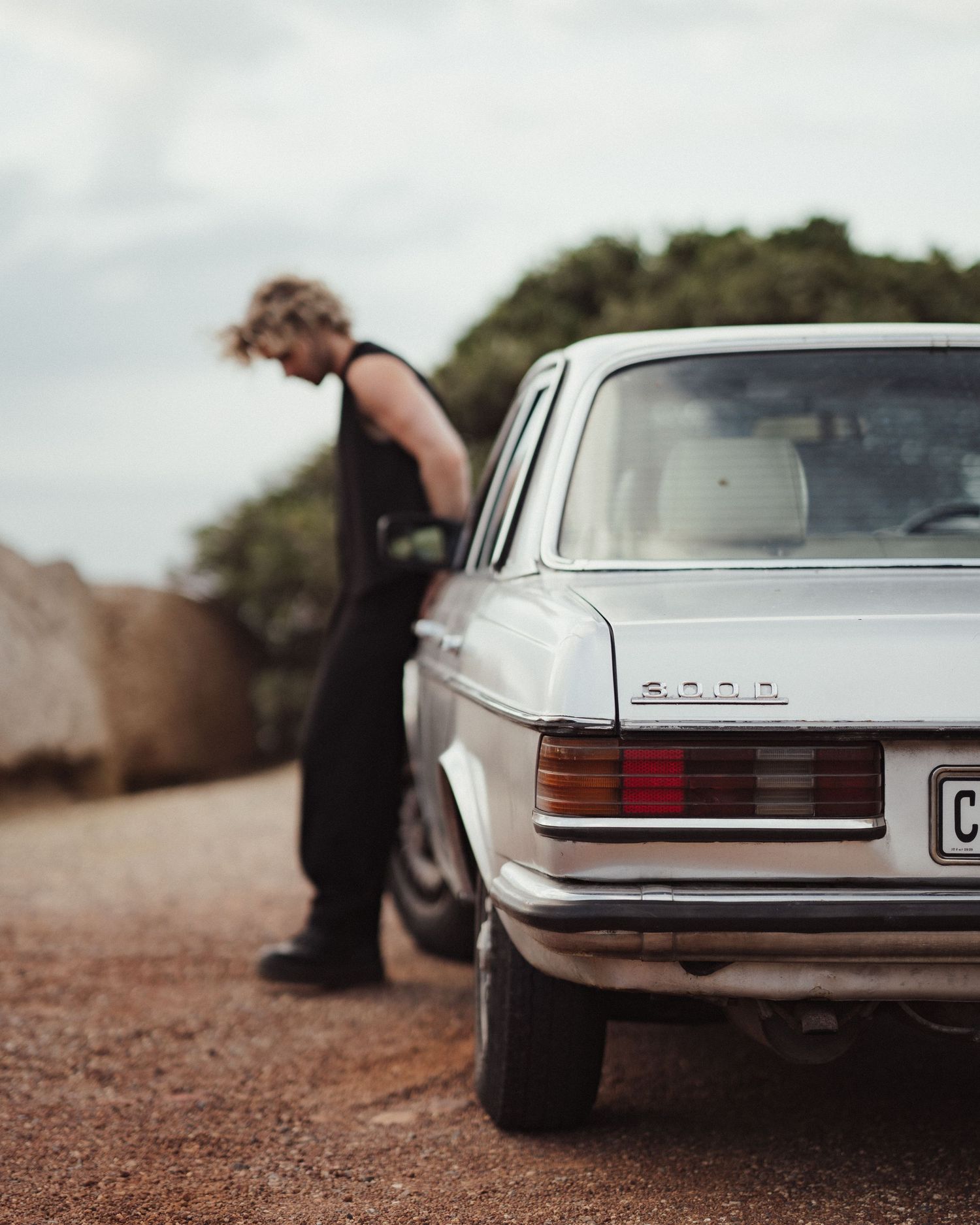 Man in black standing beside a vintage white Mercedes-Benz sedan on a dirt road.
