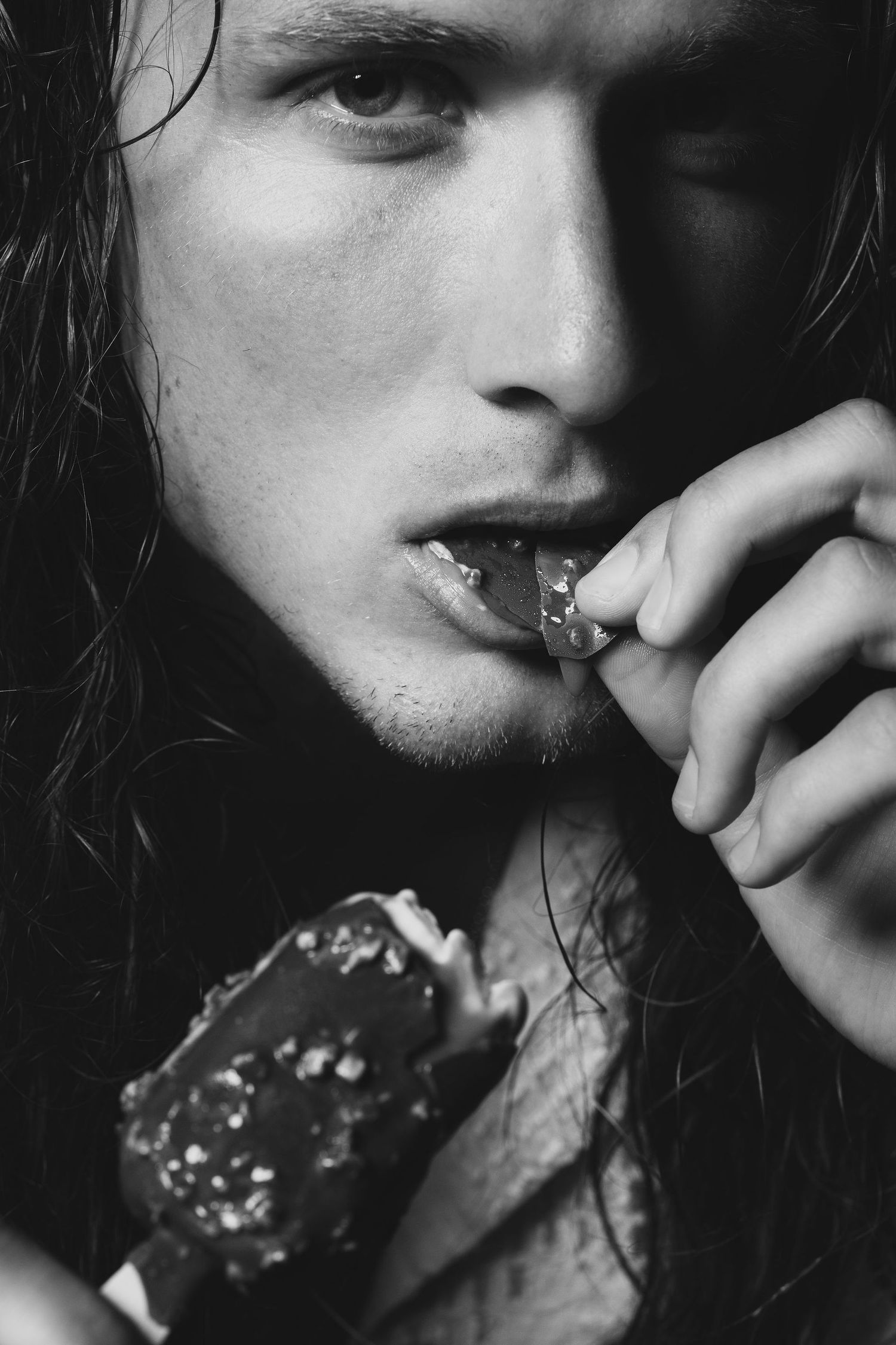 Black and white photo of a young man with long wet hair sensually eating a chocolate ice cream bar.