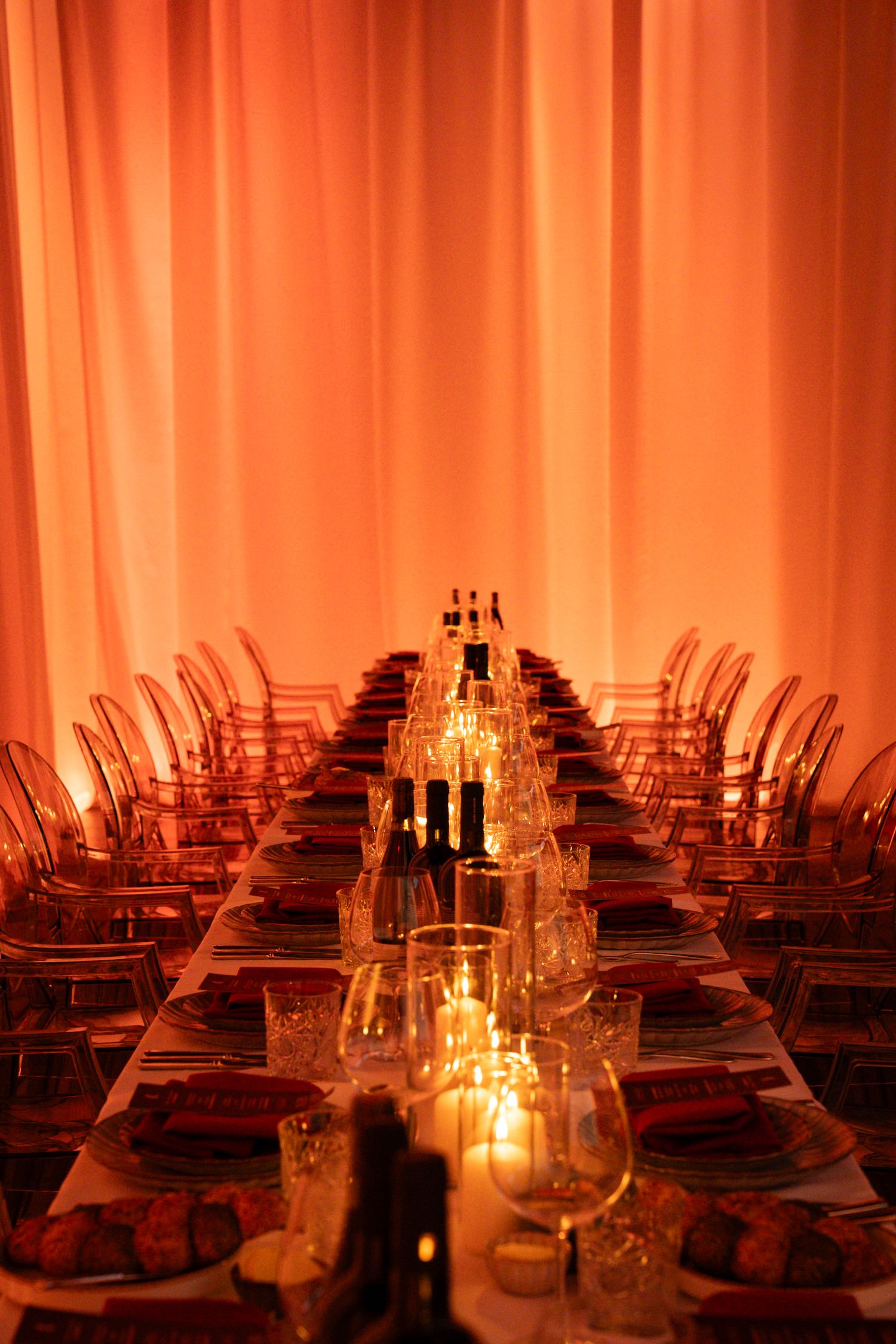 Elegant long dining table set with candles and glassware, bathed in warm orange lighting with sheer curtains backdrop.