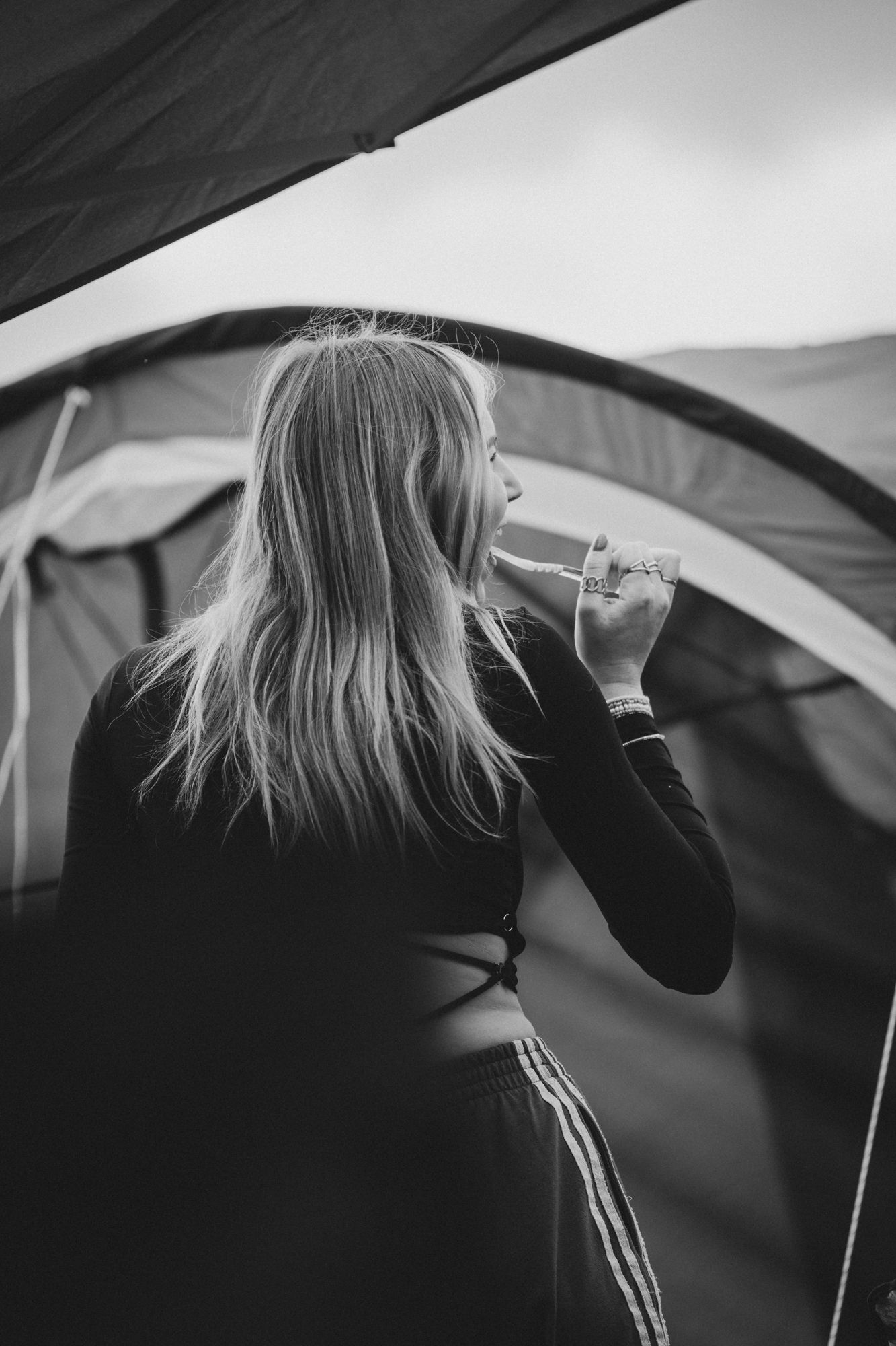 Black and white photo of a woman with long blonde hair drinking from a cup outside a camping tent.