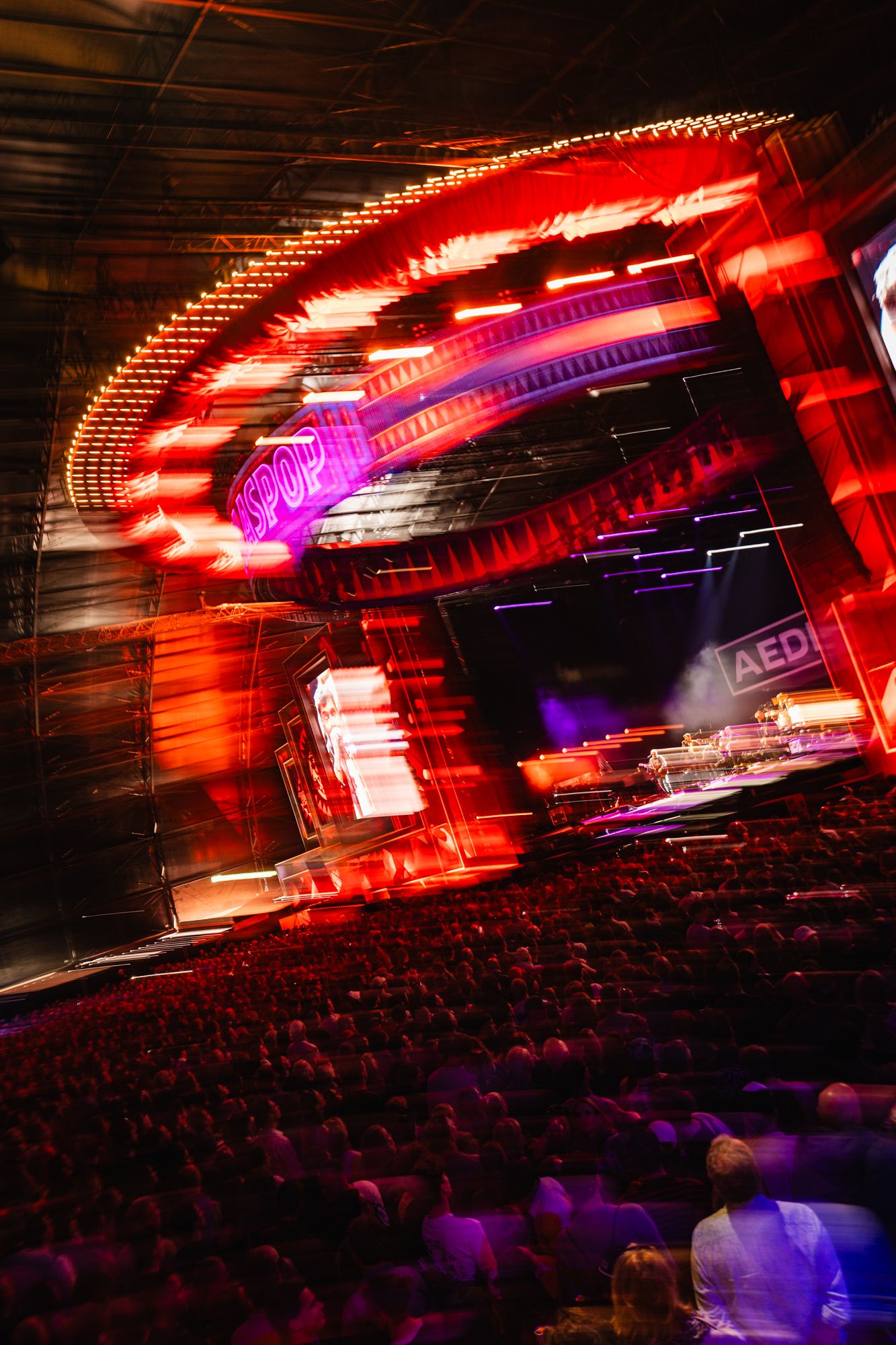 Vibrant concert stage with red neon lights and motion blur effect, packed audience in foreground at live music event.
