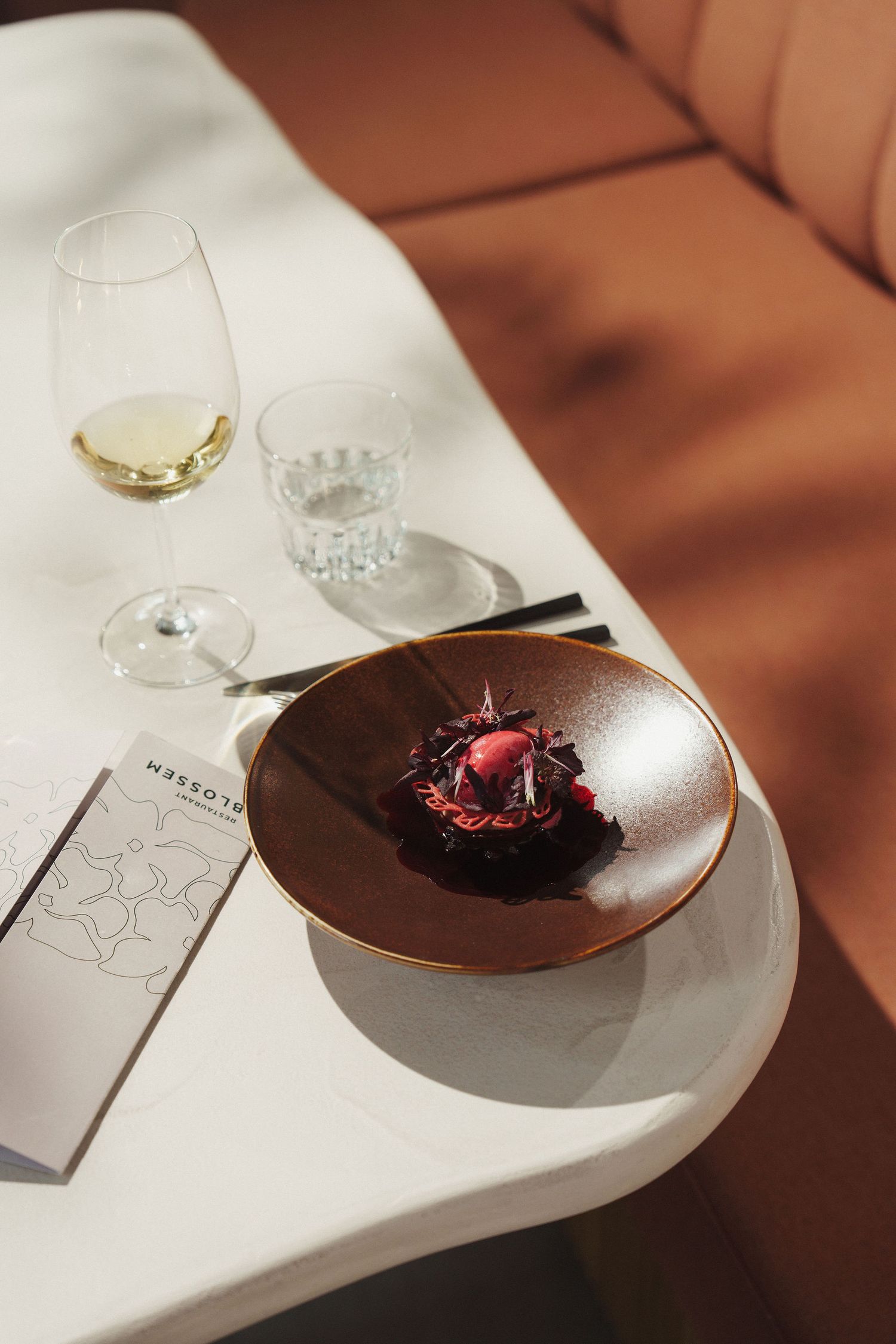 Elegant fine dining dessert on brown plate with white wine and water glass on marble table in sunlit restaurant.
