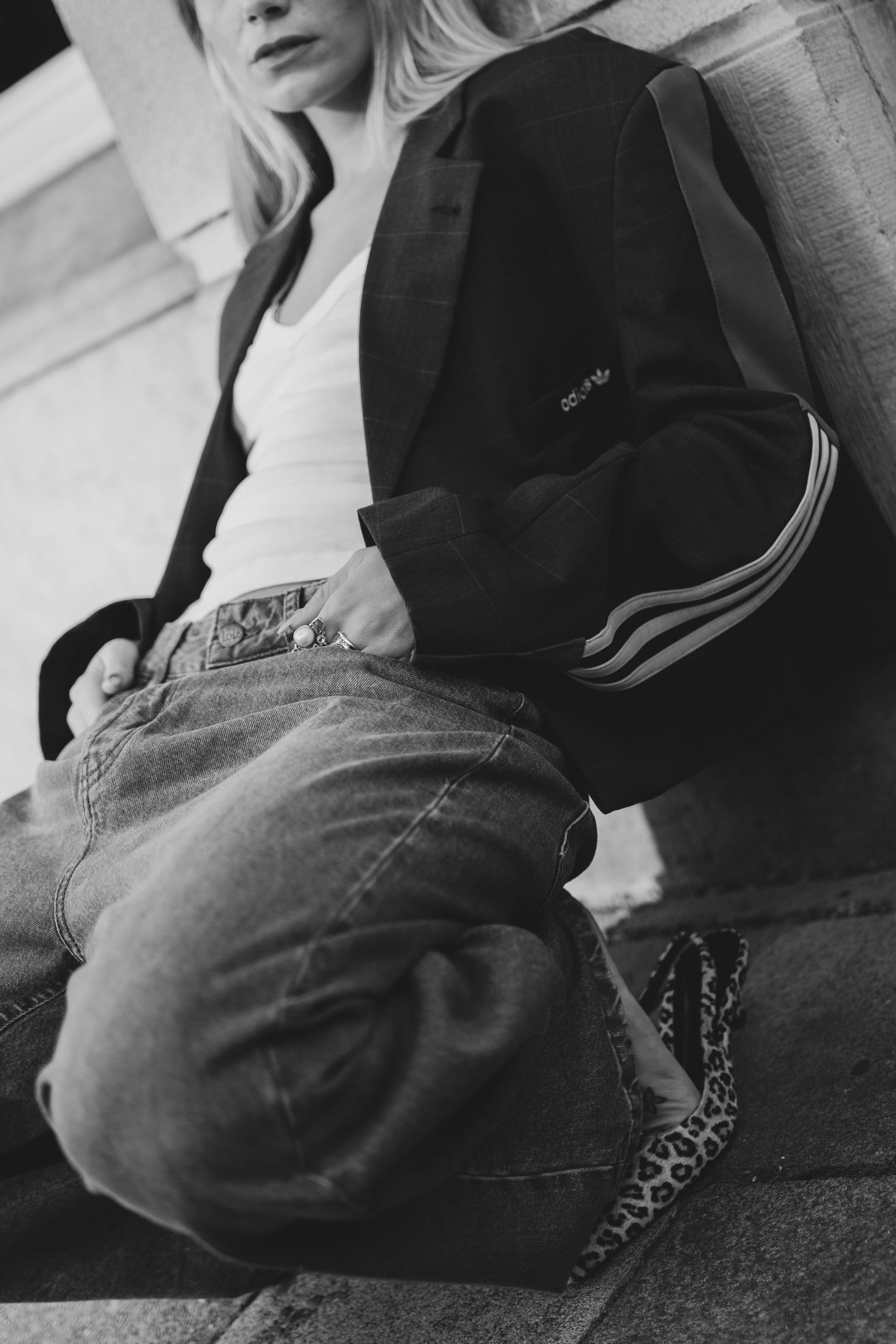 Black and white photo of a woman in an Adidas jacket, white tee, and jeans sitting against a wall.