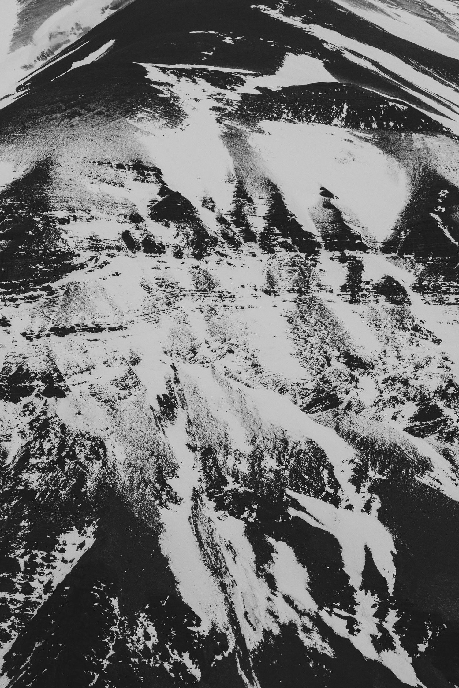 Black and white aerial photograph of a dramatic mountain landscape with snow-dusted rocky ridges and deep shadowed valleys.