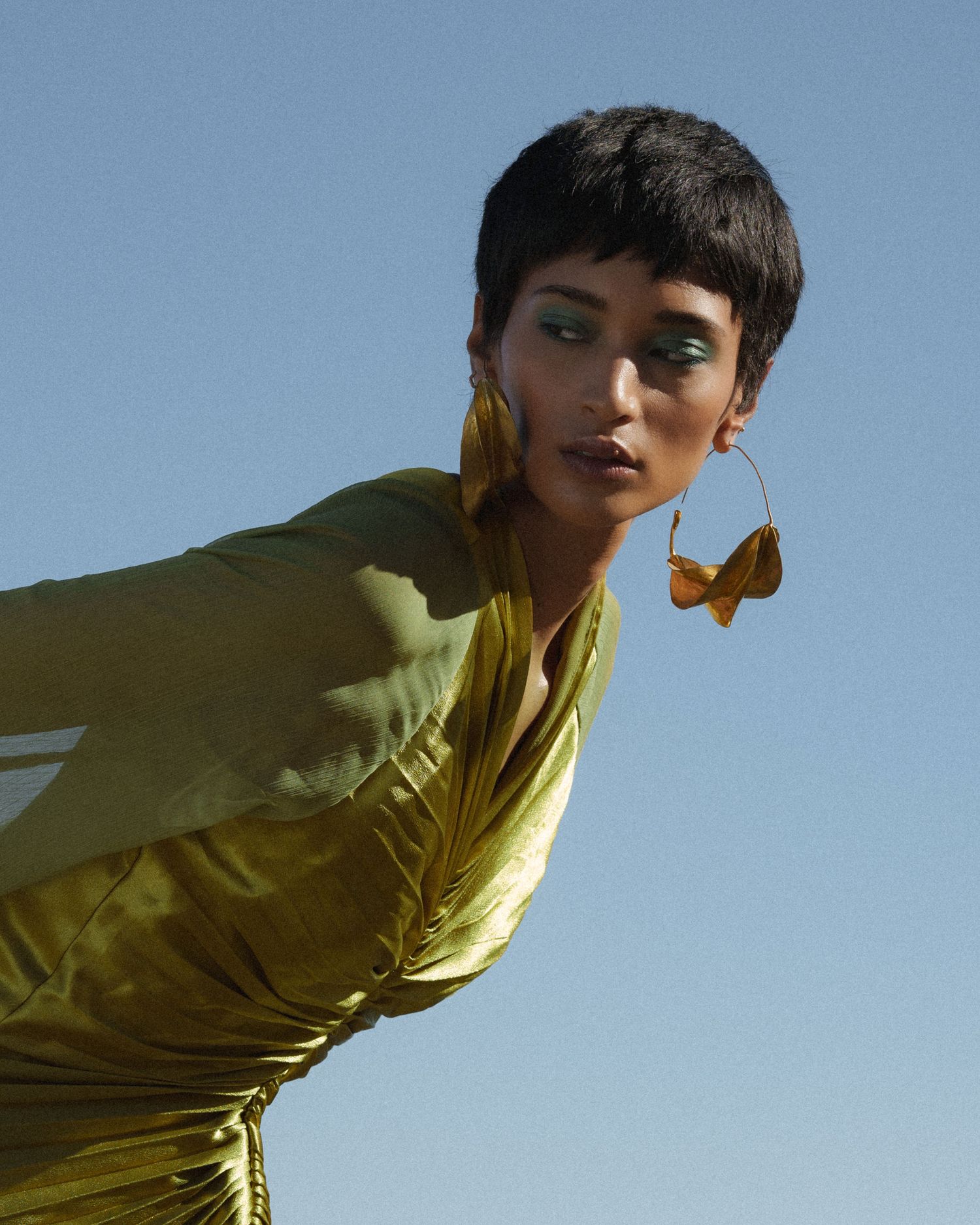 Model in gold satin dress with large hoop earrings and green eyeshadow poses against blue sky.