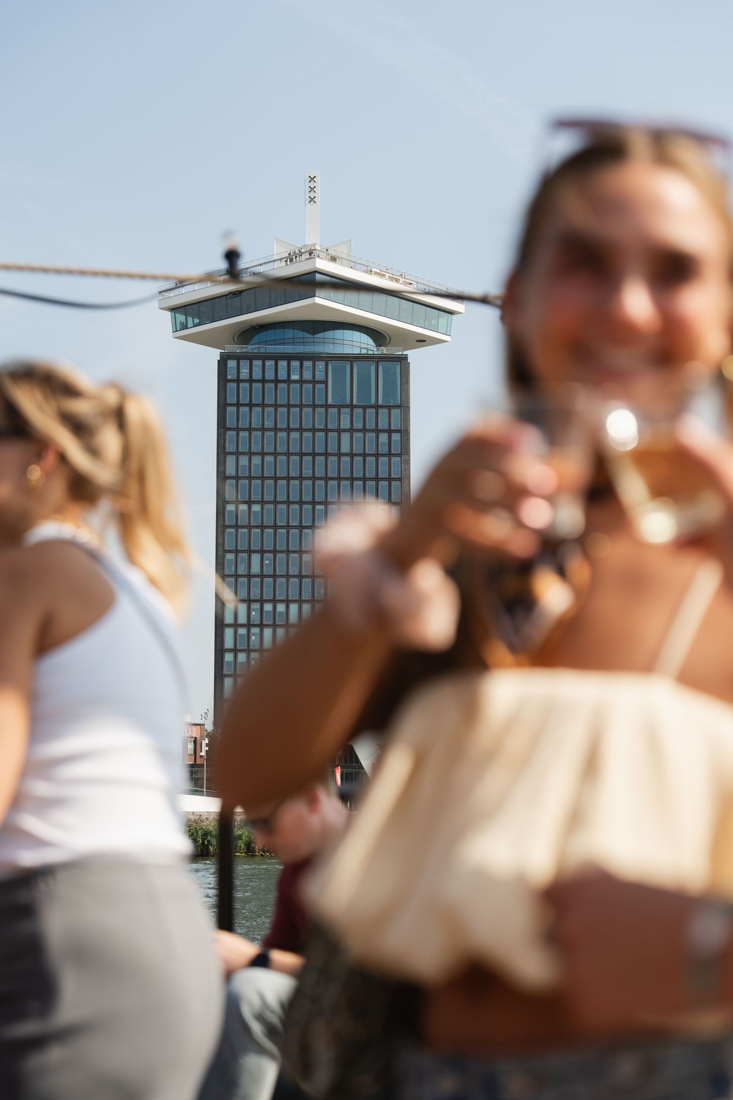 Two women enjoying drinks on a boat tour with the A'DAM Tower landmark visible in Amsterdam on a sunny day.