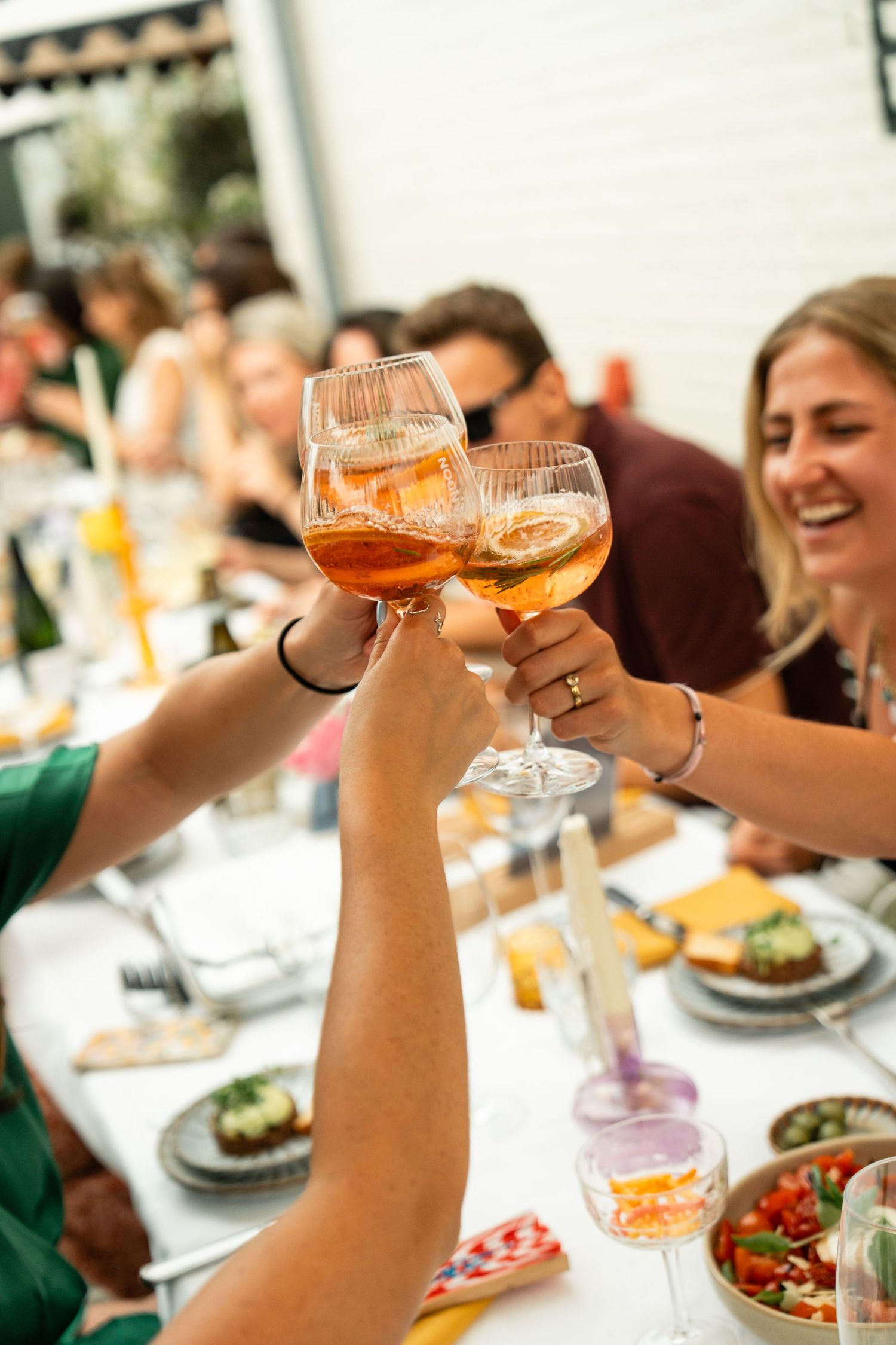Friends toasting with Aperol Spritz cocktails at a festive outdoor dinner party table with food.