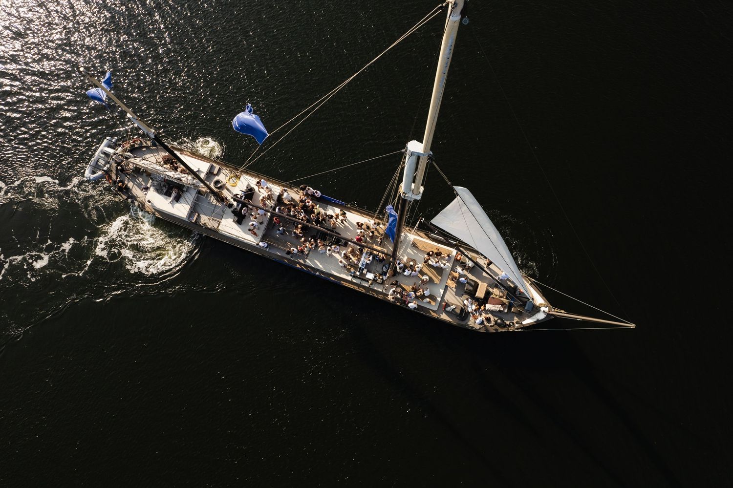 Aerial view of a sailboat with mast and rigging sailing on calm dark water with people visible on deck.