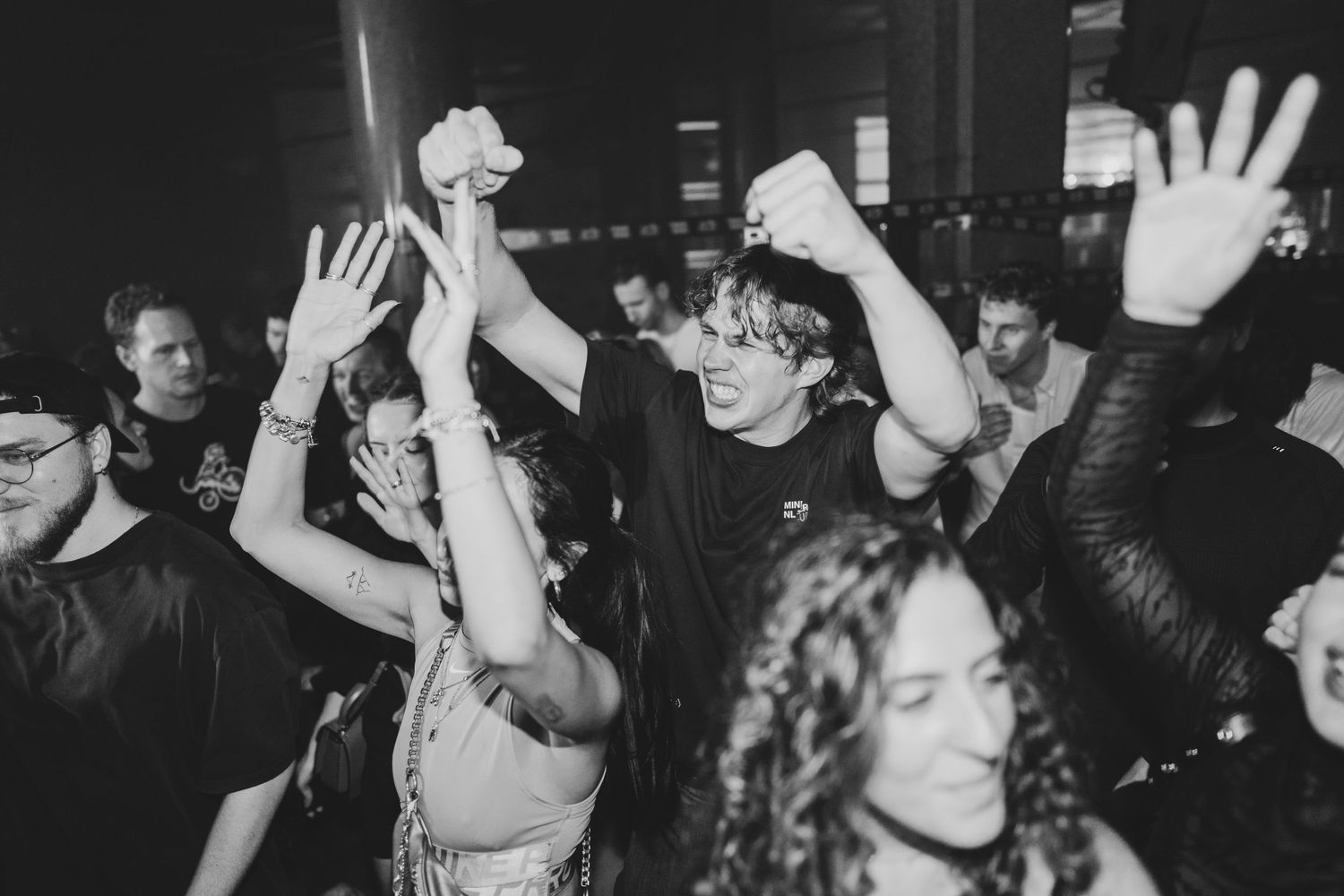 Energetic crowd dancing at a nightclub event, raising hands in excitement under dark atmospheric lighting.