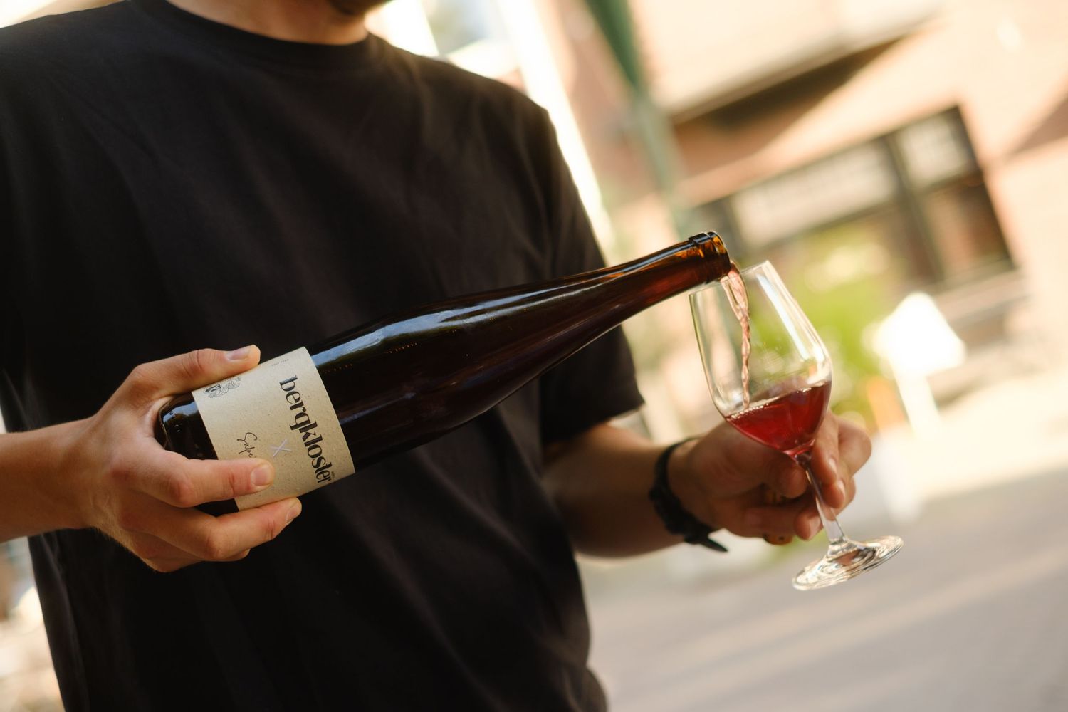 Person pouring red wine from a labeled bottle into a glass outdoors, wearing a black t-shirt.