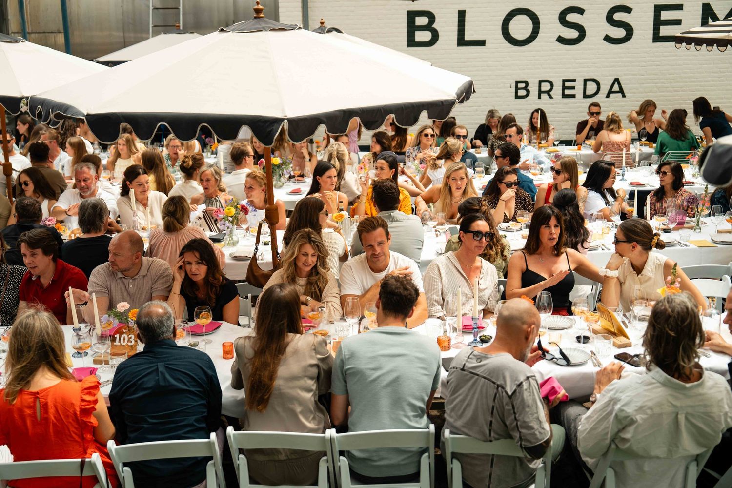 Crowded outdoor dining event at Blossom Breda with guests seated at tables under a large white umbrella.