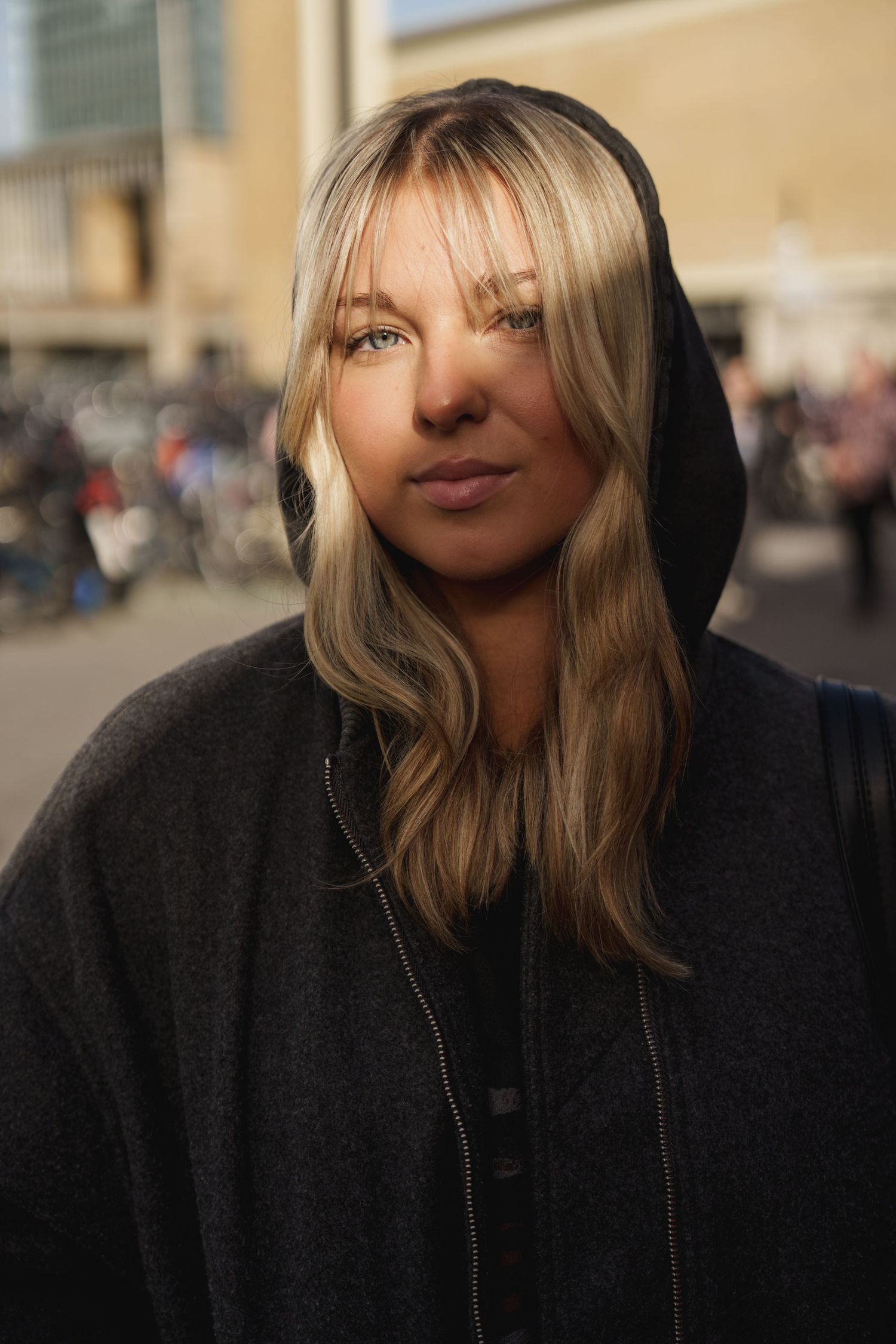 Blonde woman in a dark hoodie poses outdoors with a blurred urban background in warm sunlight.