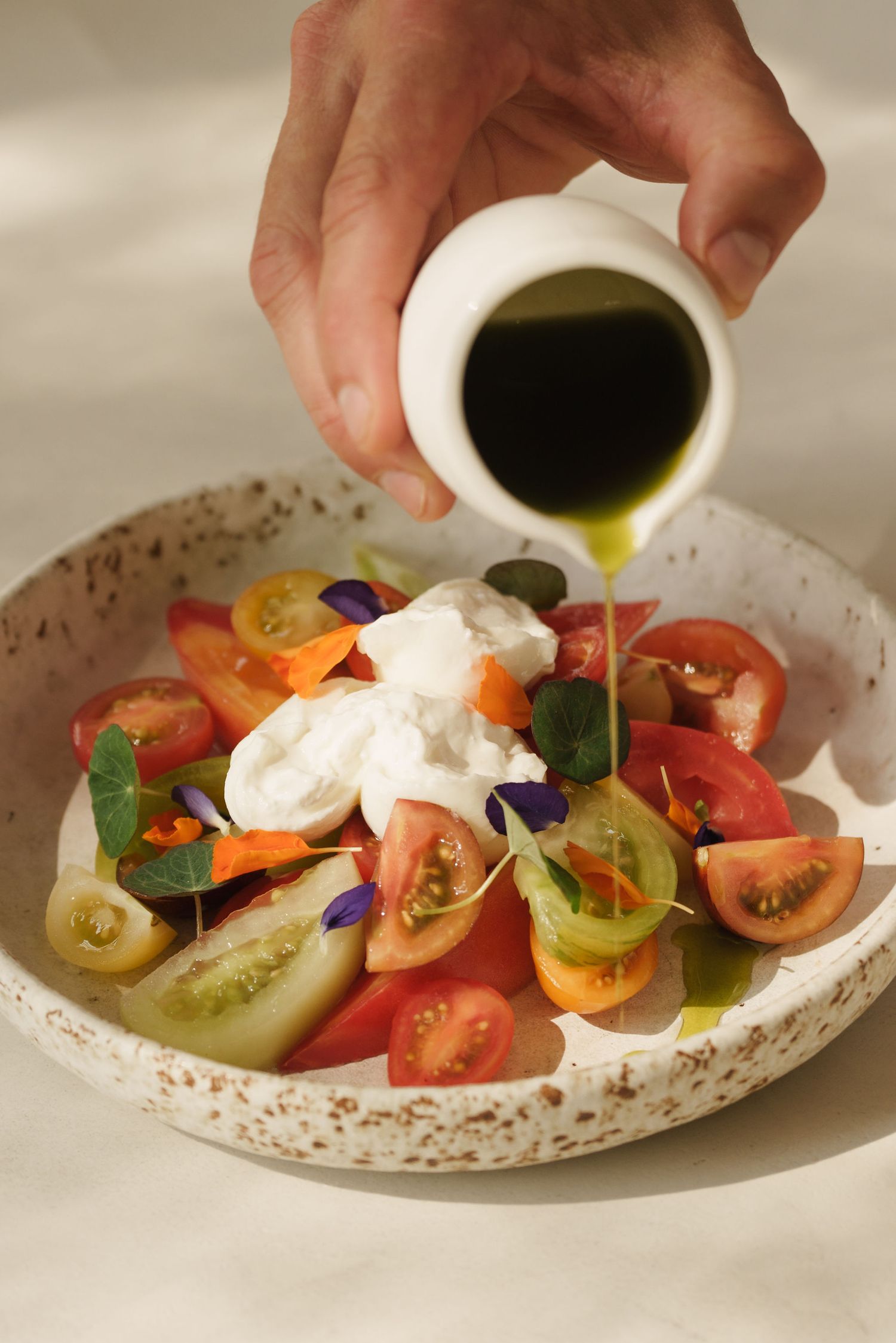 Hand pouring olive oil over fresh caprese salad with heirloom tomatoes, burrata, and edible flowers in ceramic bowl.