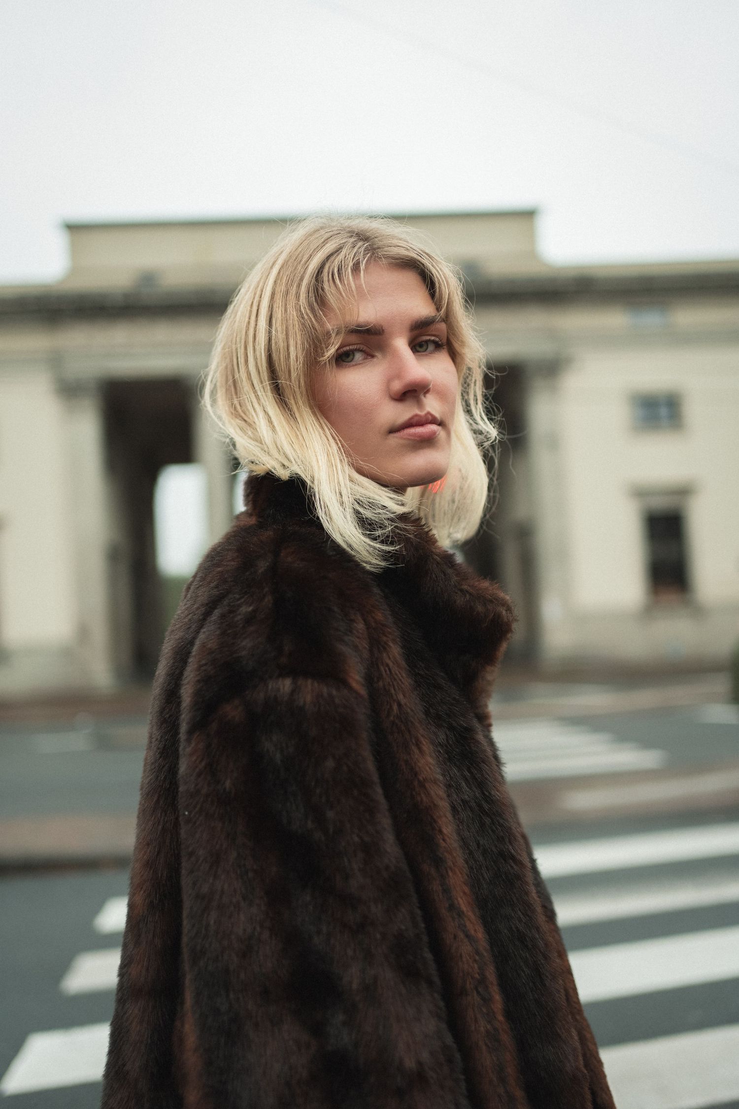 Blonde woman in dark brown fur coat standing near a crosswalk with a classical building in the background.