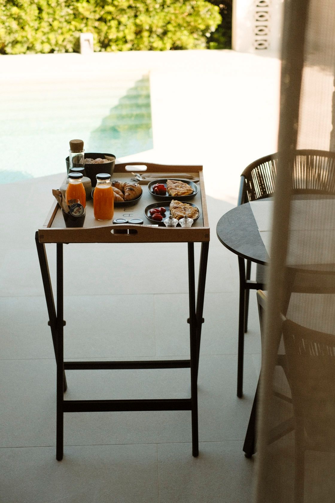 Wooden breakfast tray with orange juice, pastries, and snacks on a folding table beside a pool.
