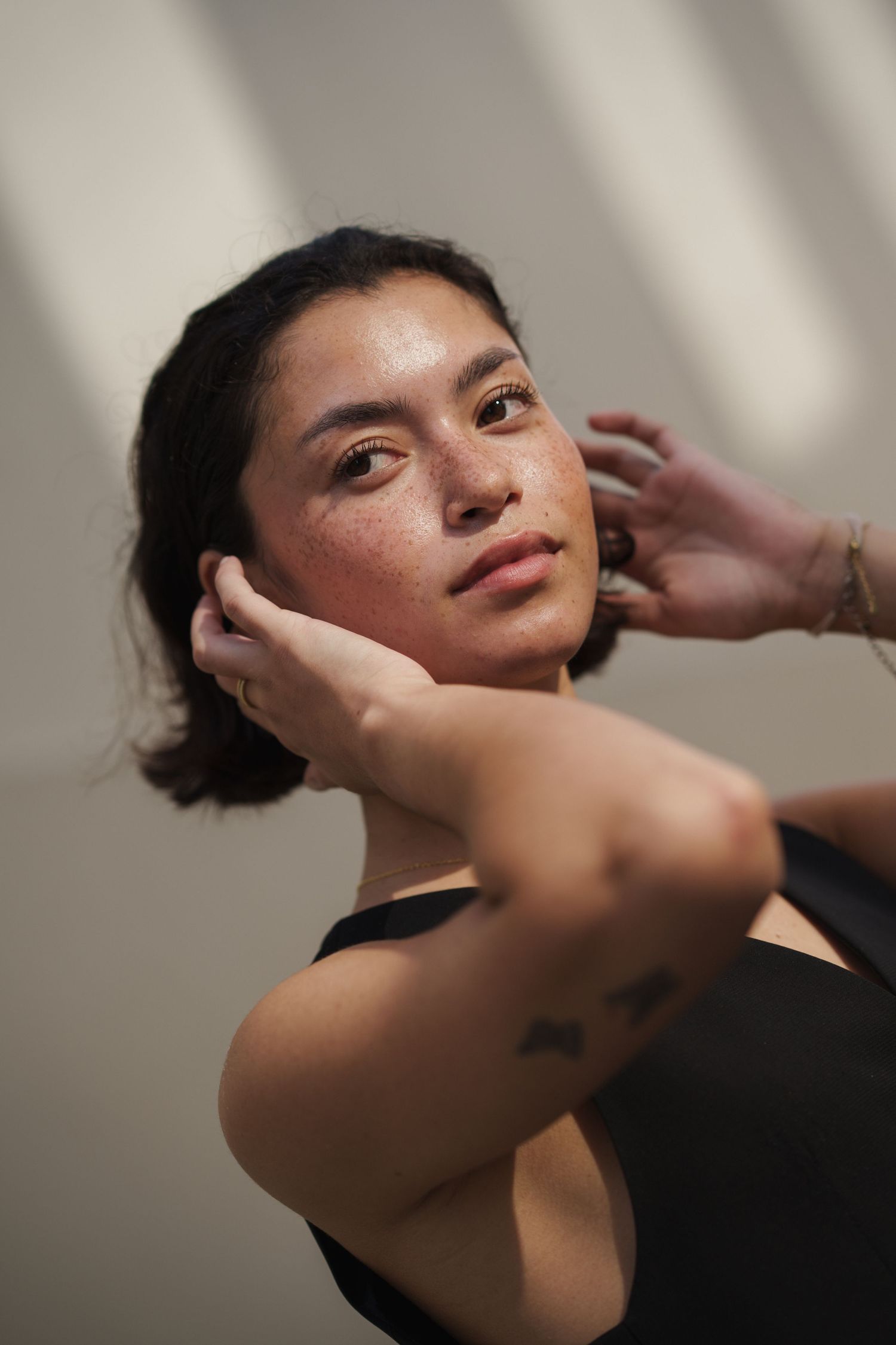 Young woman with freckles and gold eyeshadow poses gracefully with hands near face, wearing black top with arm tattoo.