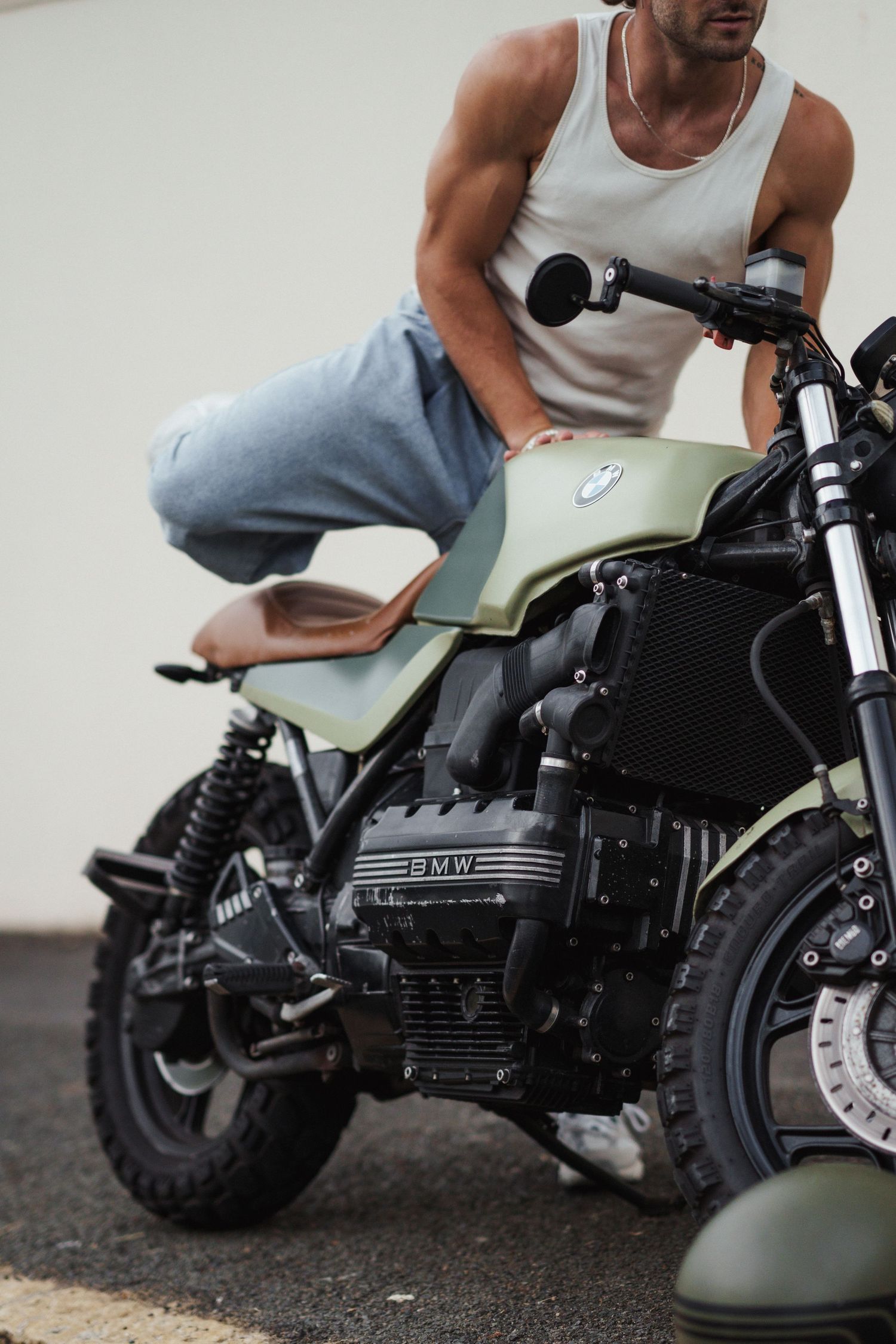 Man in white tank top and jeans sitting on a custom matte green BMW motorcycle outdoors.