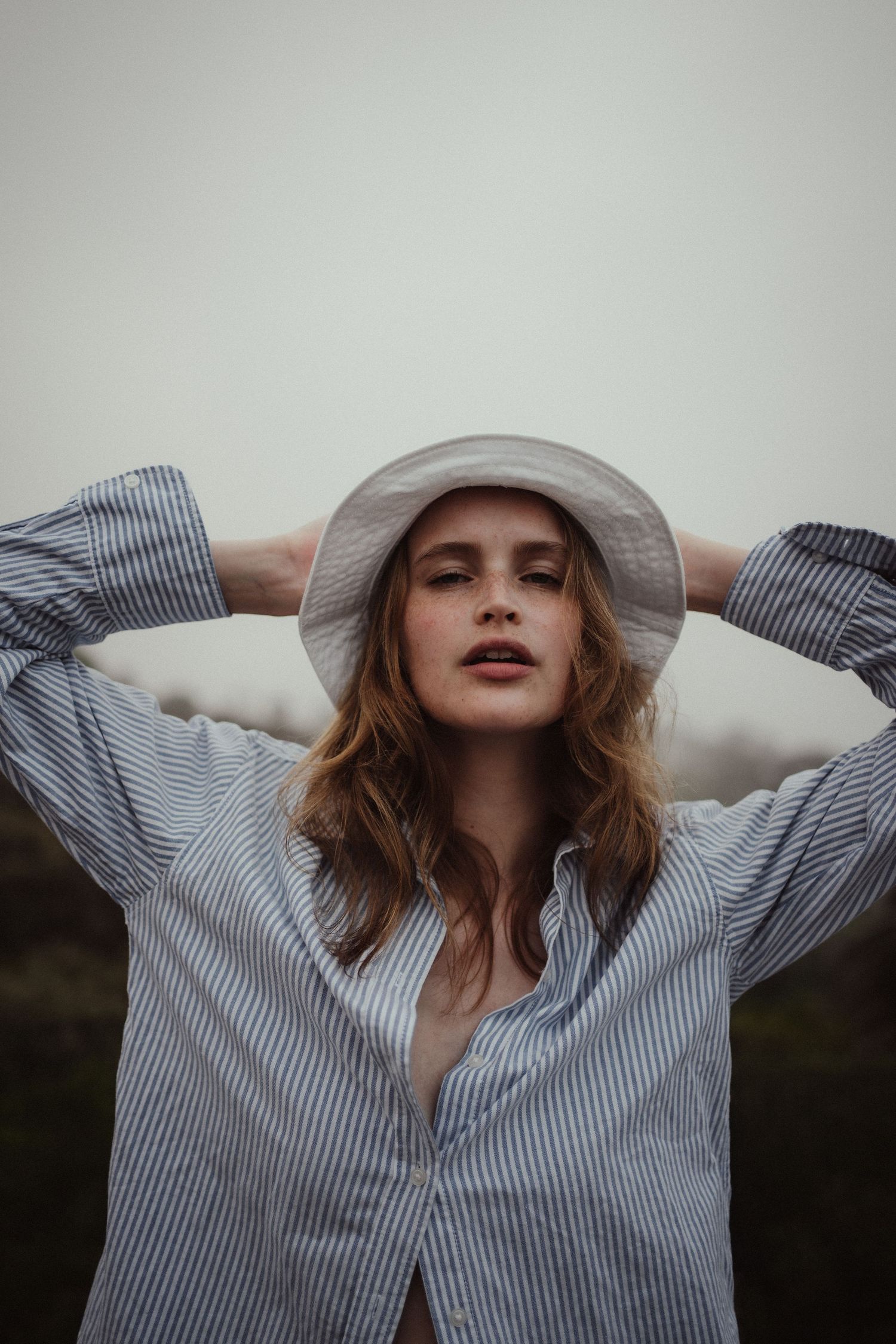 Young woman in striped shirt holding white hat on head, posing against overcast sky.
