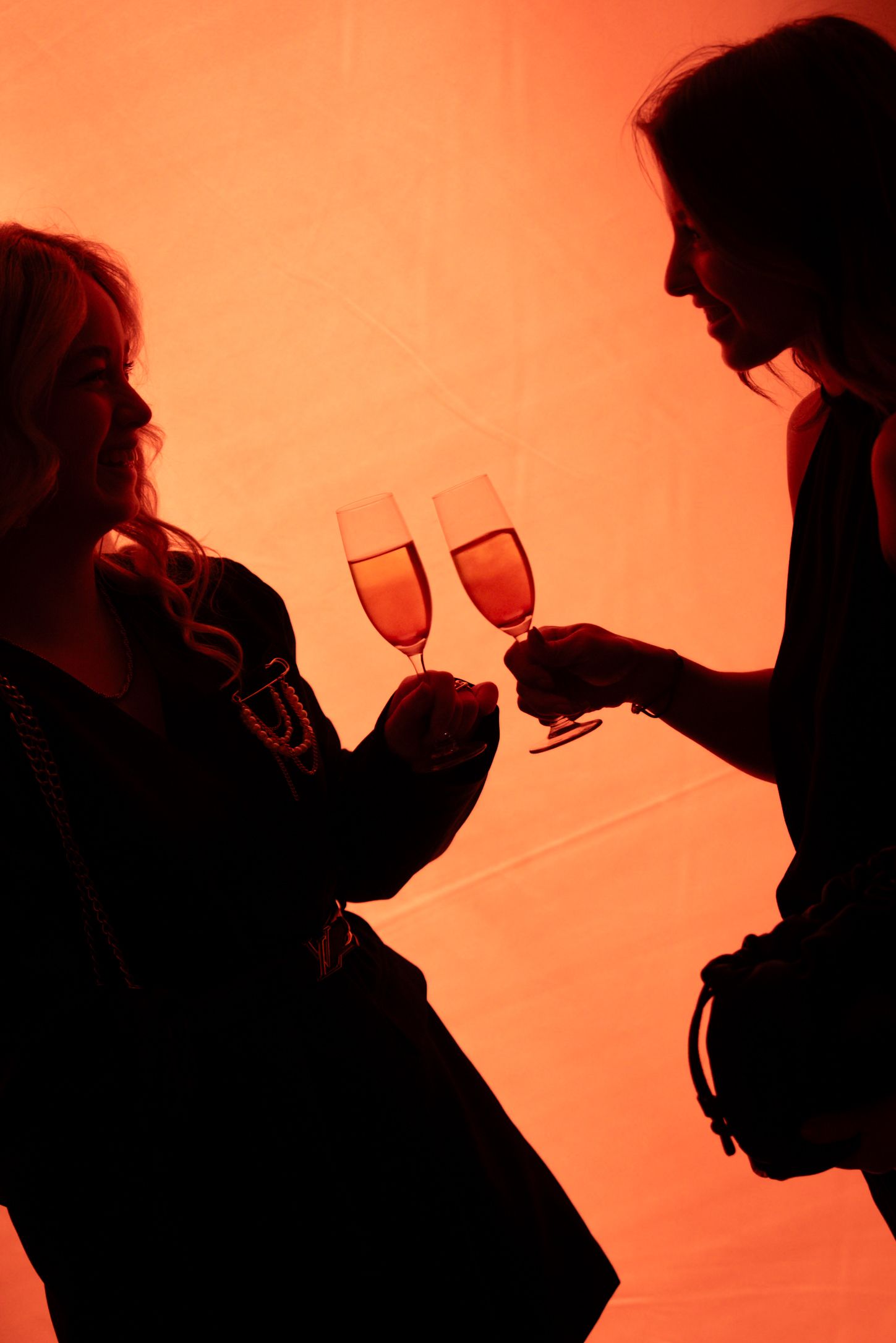 Two women clink champagne flutes in a warm red-orange silhouette glow.