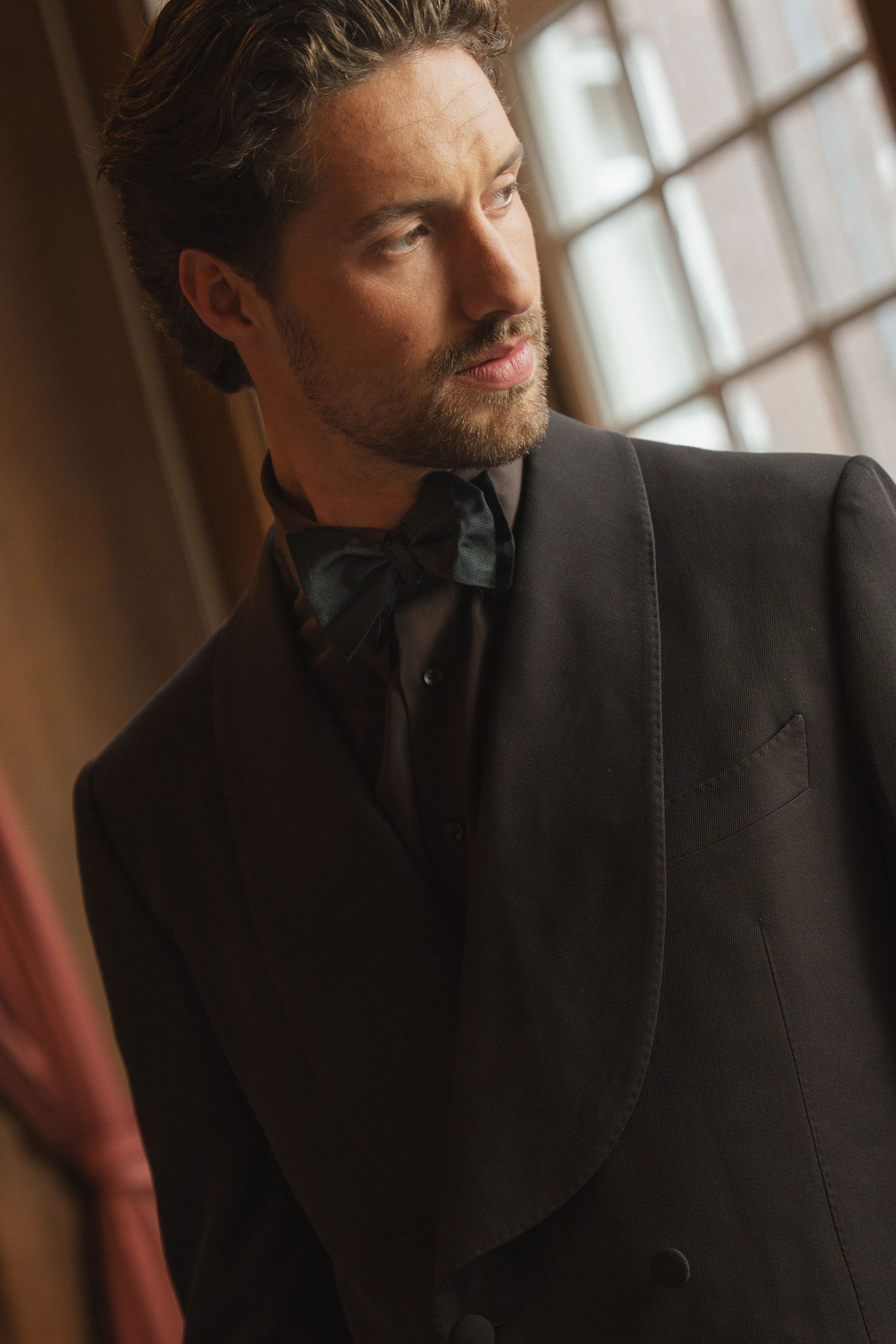 Elegant man in black tuxedo with bow tie gazing sideways near a window in formal attire.
