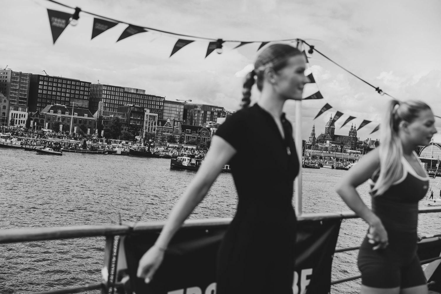 Two women stand on a dock with colorful bunting flags and a harbor cityscape in black and white.