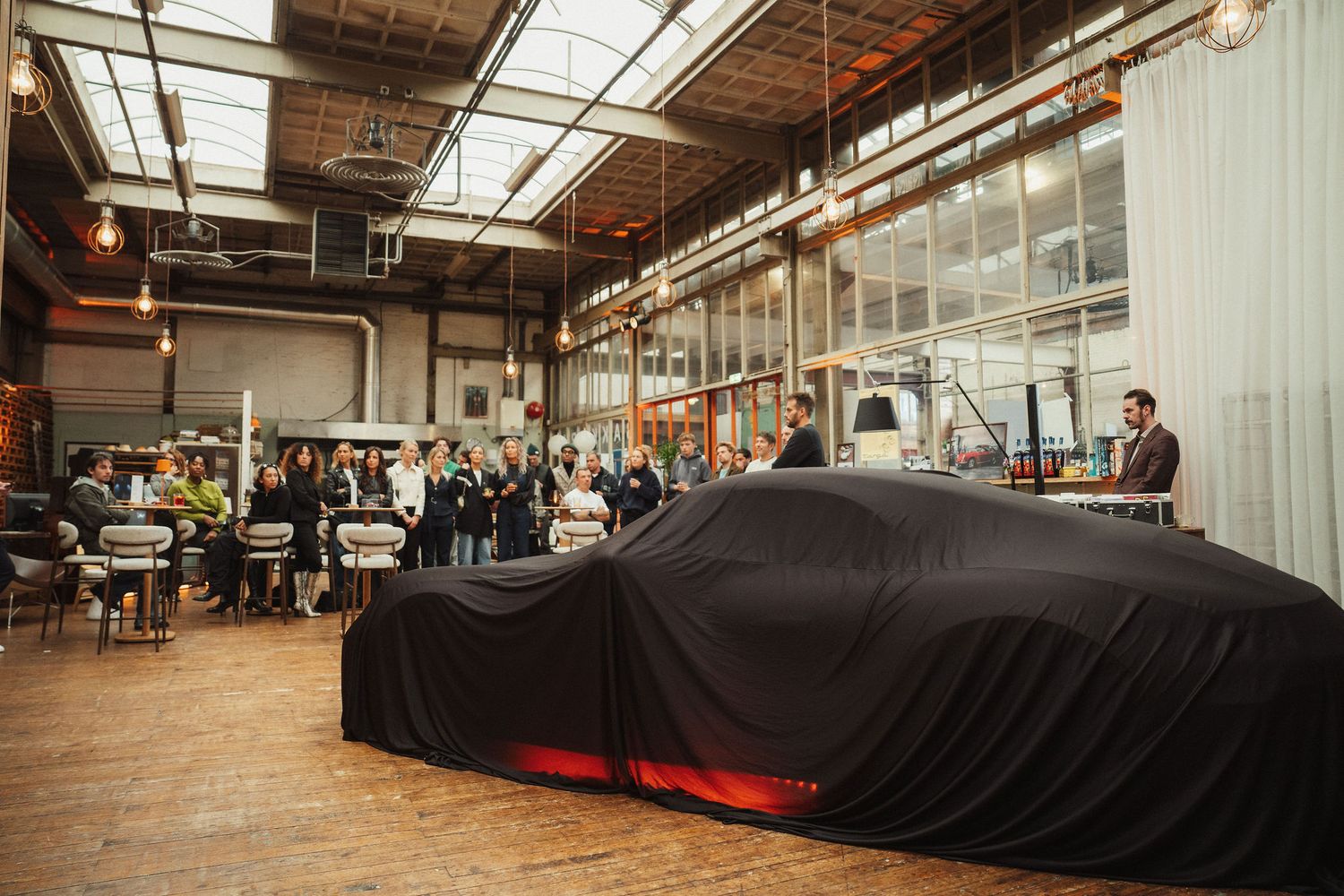 A sleek car covered in black cloth unveiled at an industrial warehouse event with guests gathered around.
