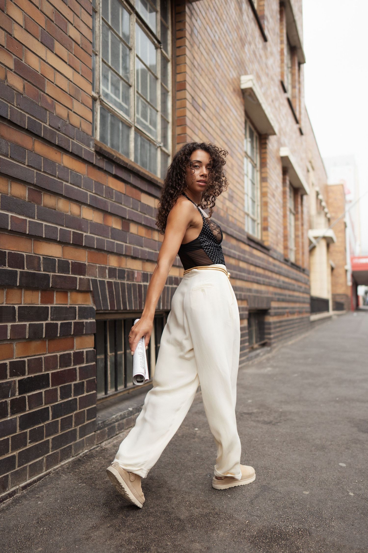 A stylish woman in a black lace top and white wide-leg pants walks along a brick building sidewalk.