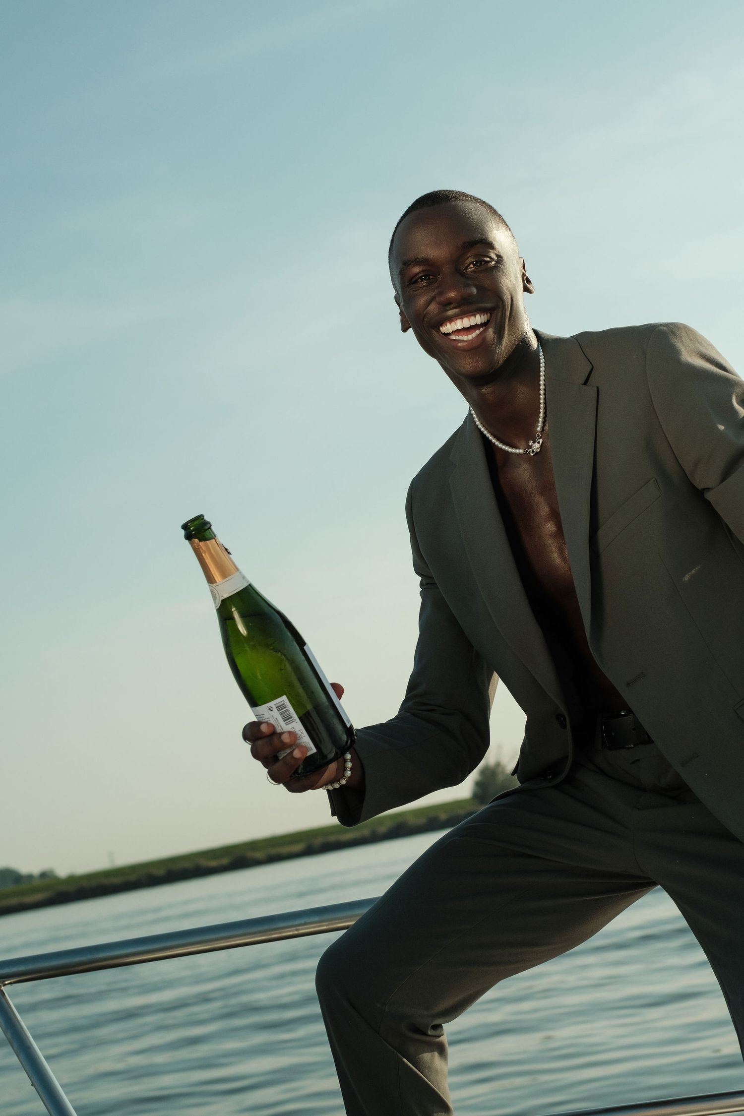 Smiling Black man in gray suit holding champagne bottle on yacht deck with blue sky background.