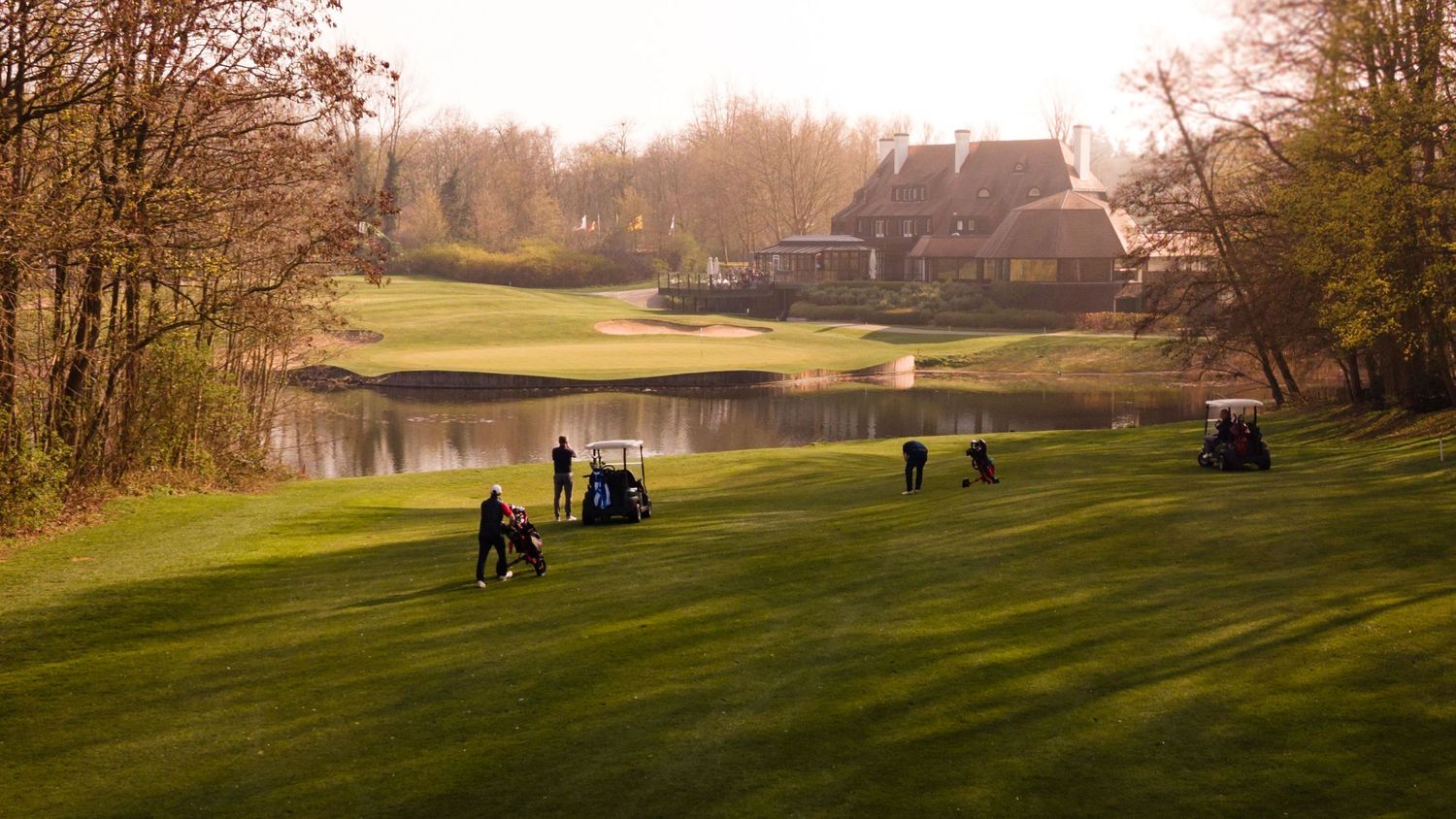 Golfers playing on a lush green golf course with a pond and rustic clubhouse in the background at sunset.