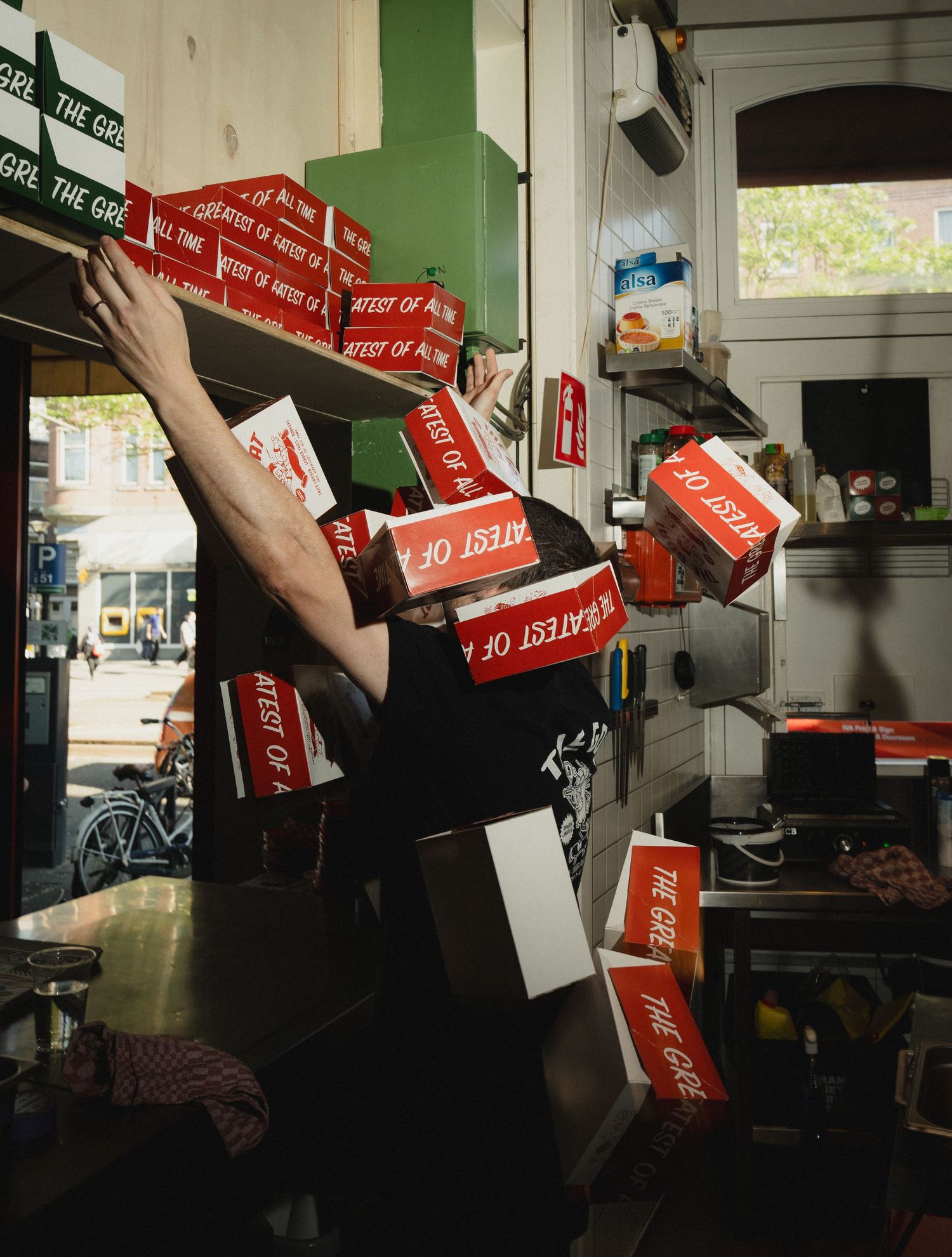Worker in black shirt juggling falling red Greatest Of All Time burger boxes in a busy restaurant kitchen.