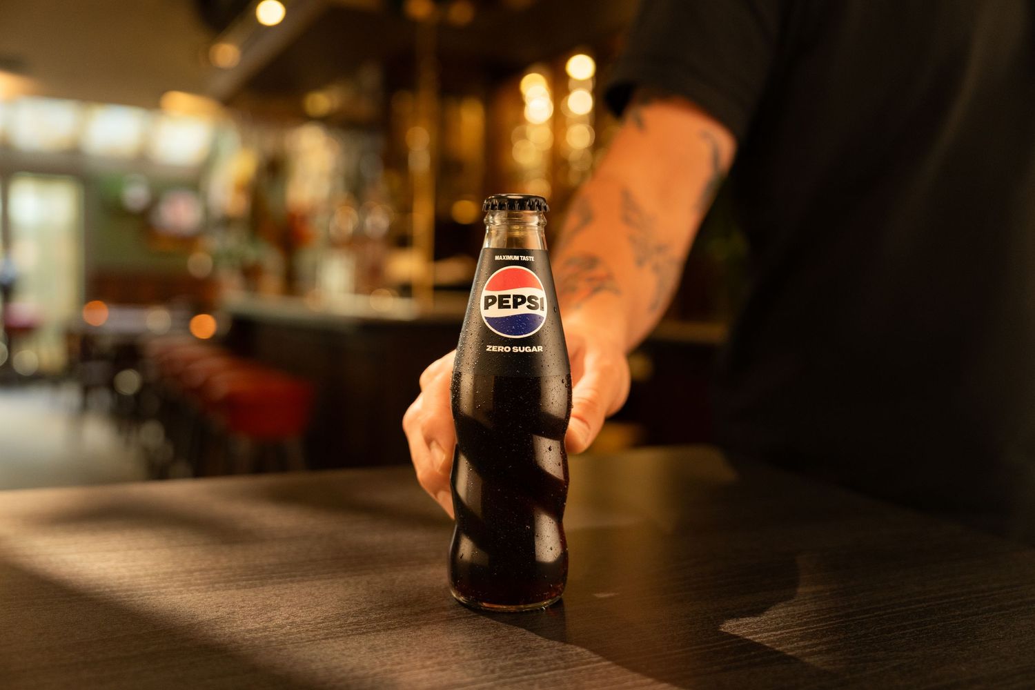 A tattooed hand places a dark Pepsi glass bottle on a wooden bar counter in a warmly lit restaurant.